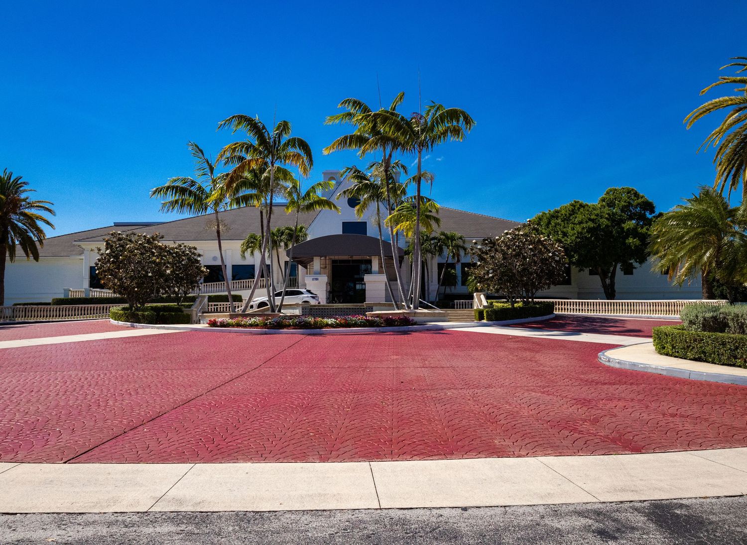 Grand Florida community clubhouse with red brick circular driveway, tall palm trees, and blue sky.