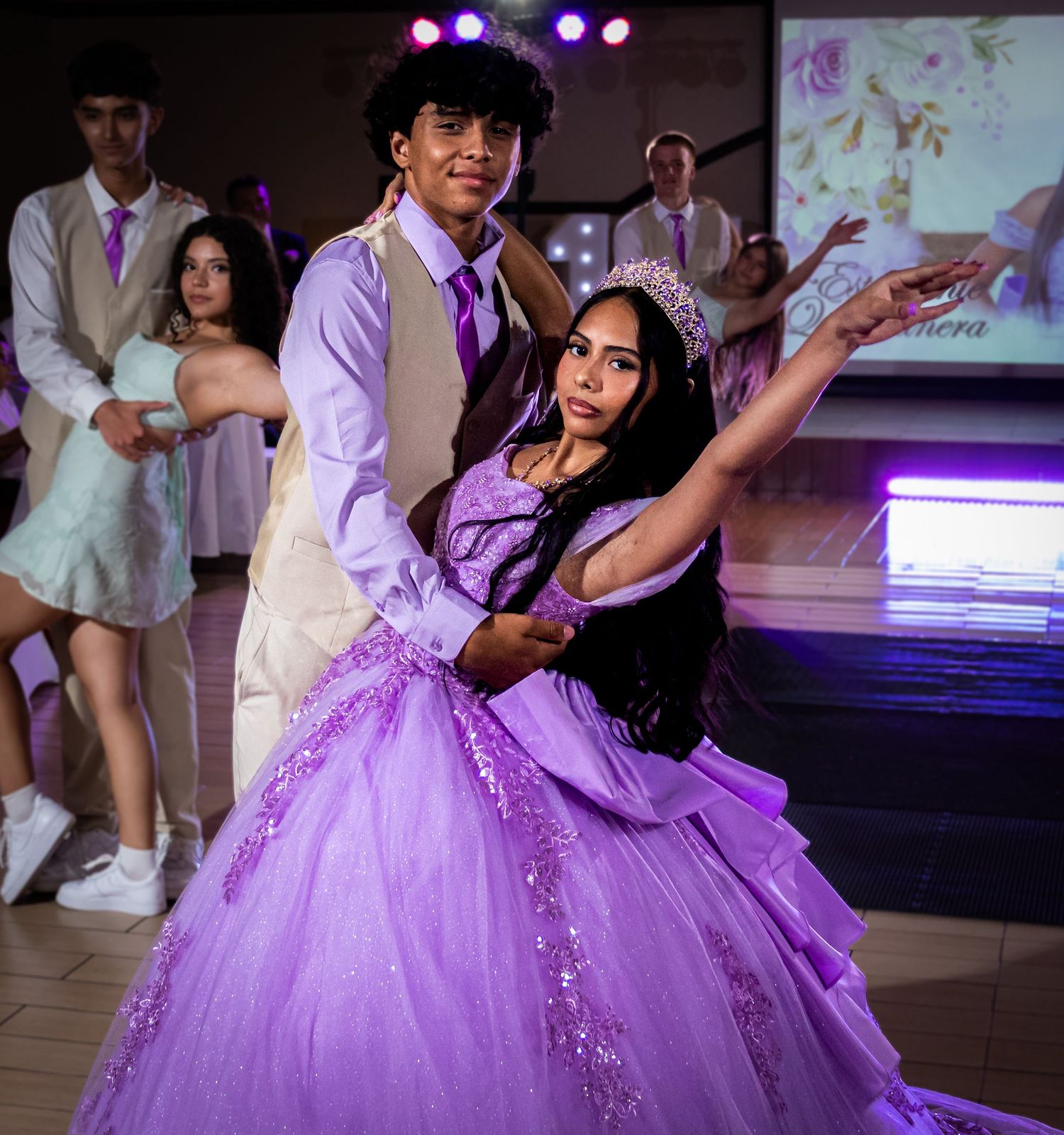 A young couple dances at a quinceañera, the girl wearing a sparkling purple ball gown and tiara under festive lights.