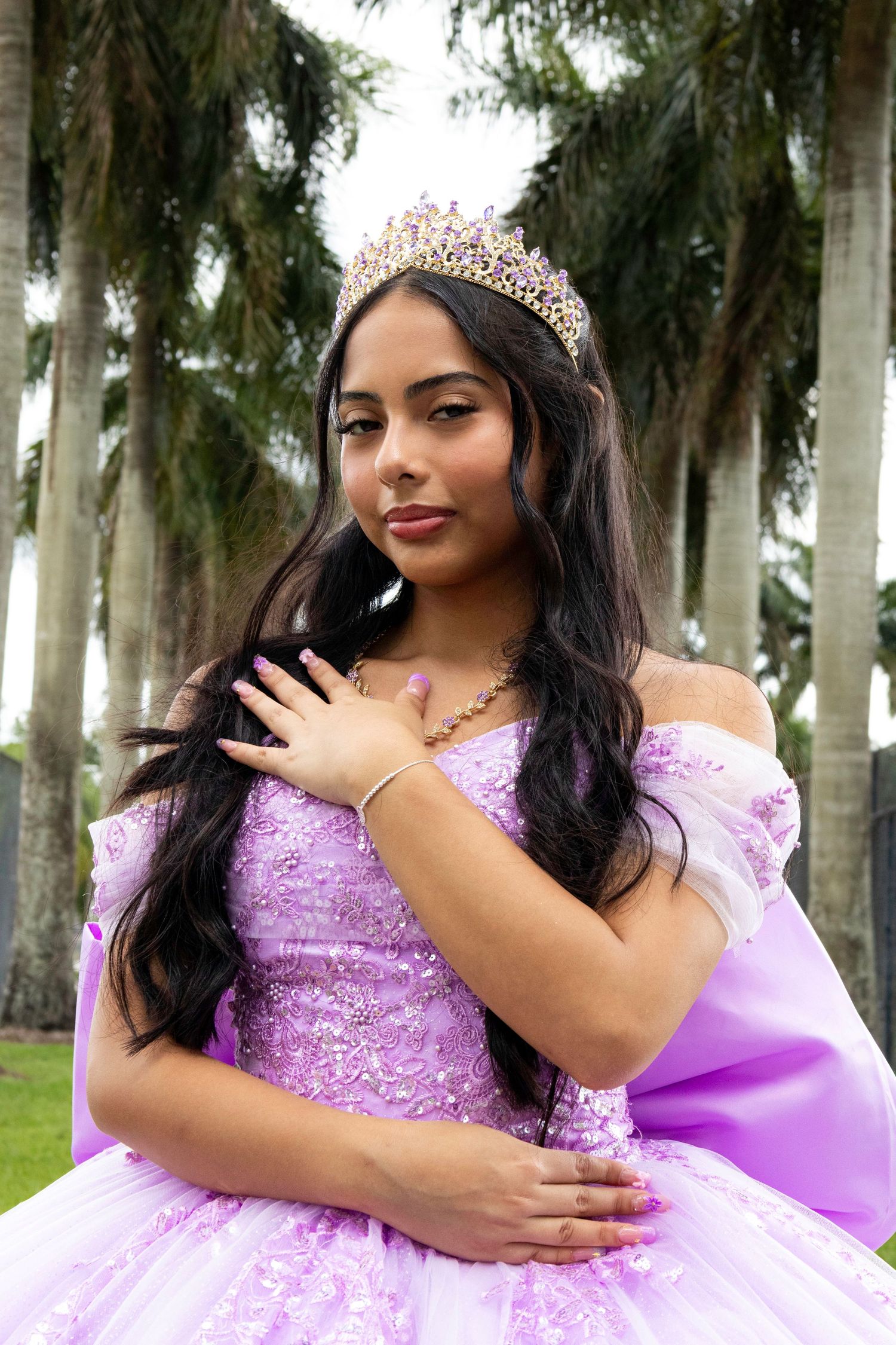 Young woman in lavender quinceañera gown and tiara poses gracefully outdoors among palm trees.