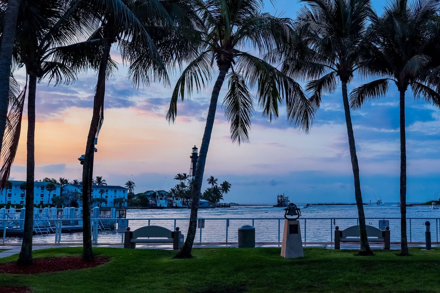 Tropical waterfront park at dusk with silhouetted palm trees, benches, and a colorful pastel sunset over calm water.