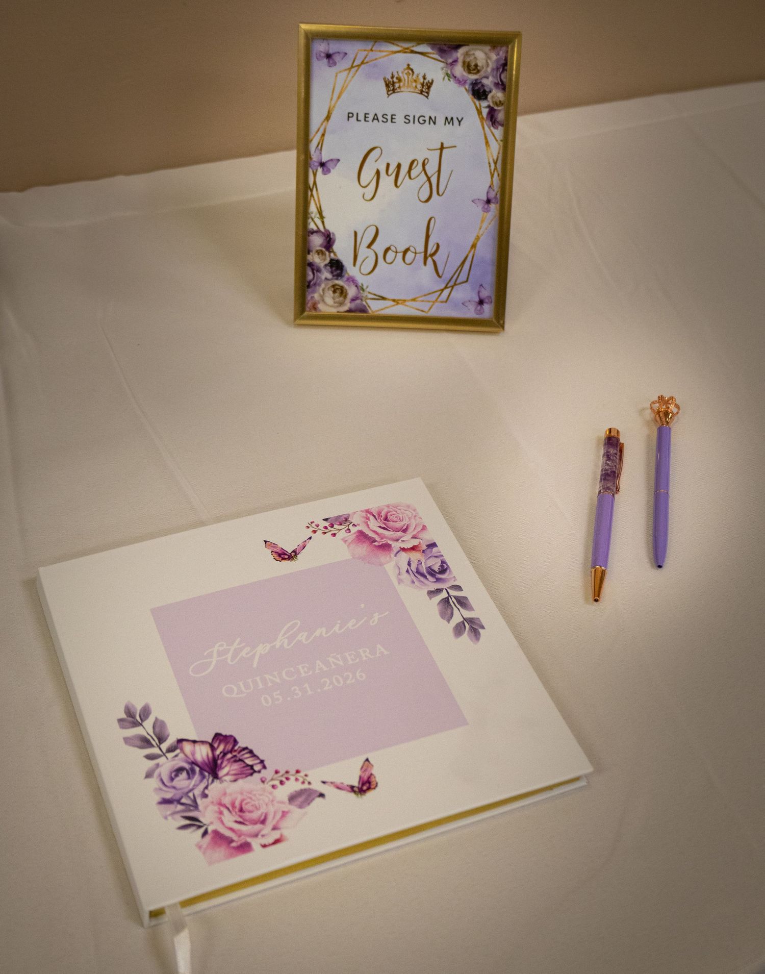 Elegant Quinceañera guest book with purple floral design, gold-framed sign, and decorative pens on white table.