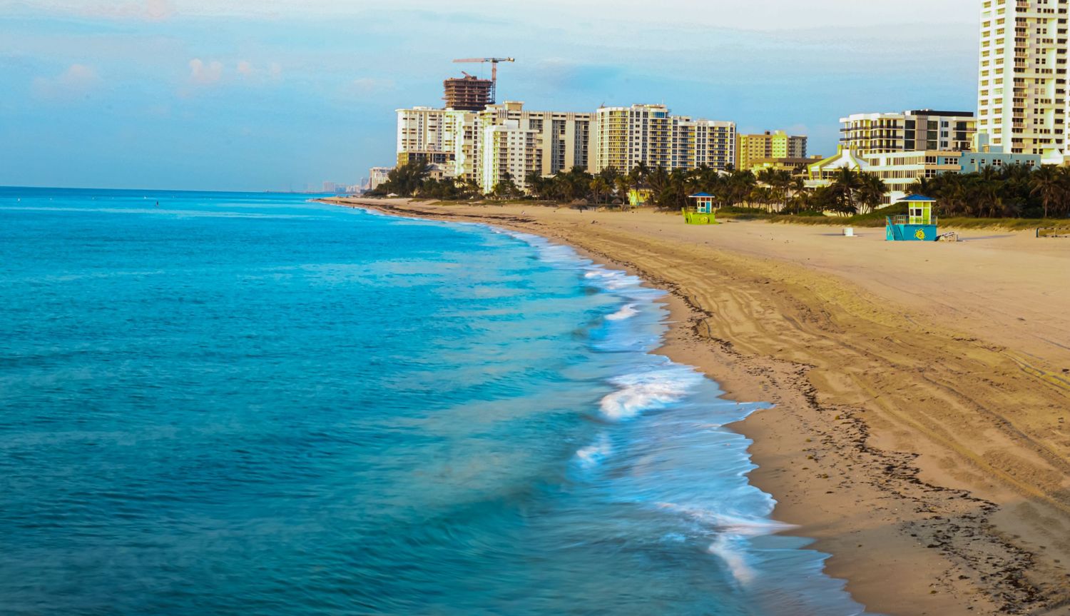 Aerial view of a pristine Florida beach with turquoise ocean waves, white sand, and luxury high-rise condos at sunrise.
