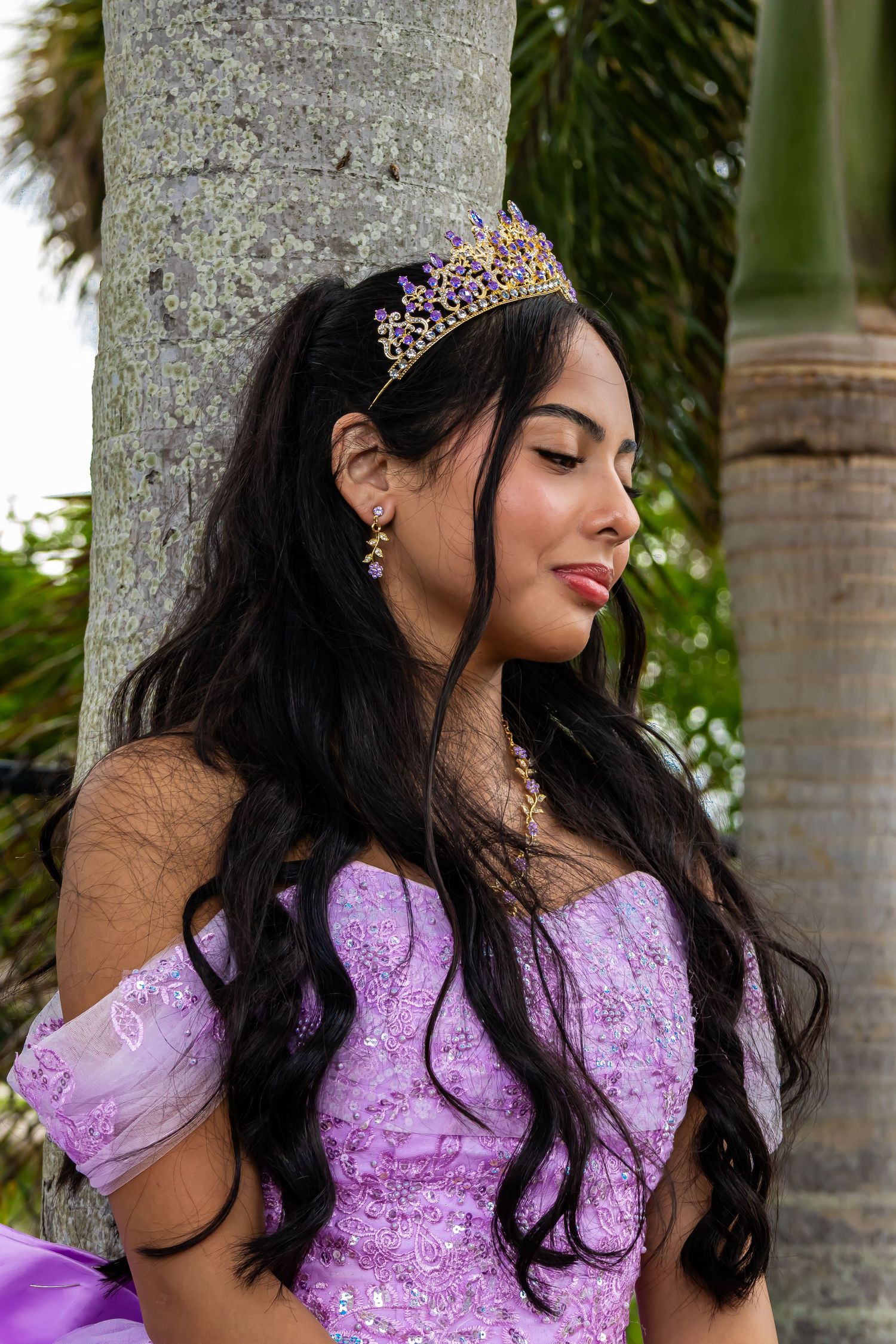 Young Latina woman in lavender quinceañera gown and purple tiara poses gracefully against a palm tree outdoors.