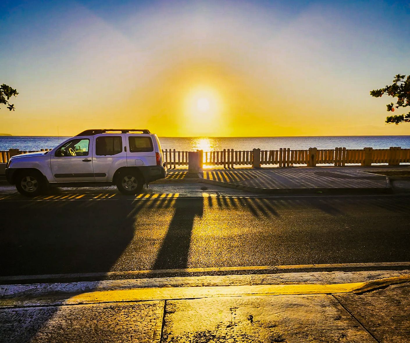 White SUV parked on a coastal road with a brilliant golden sunset over the ocean in the background.