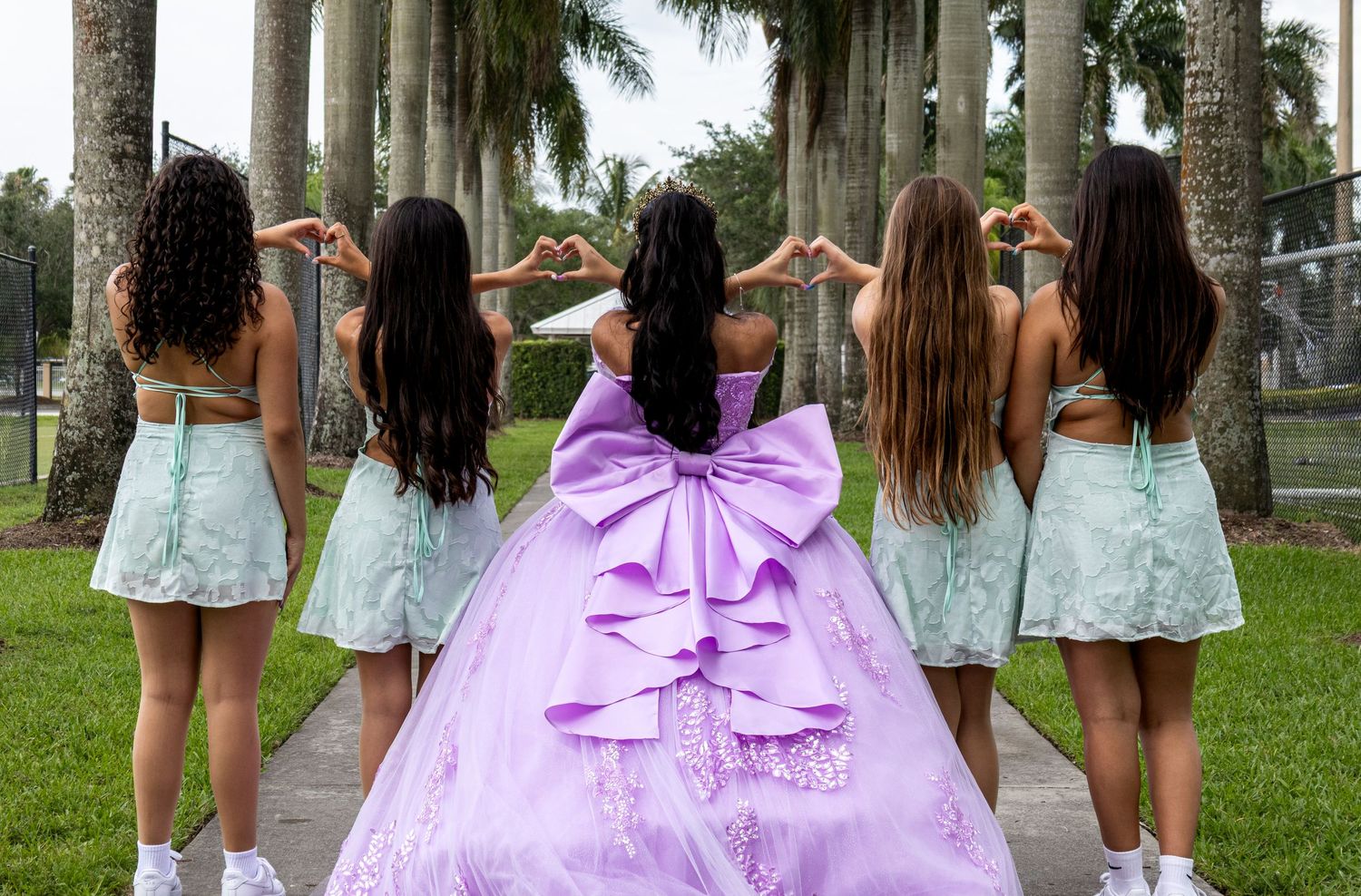 Girl in lavender quinceañera gown with large bow poses with five bridesmaids in mint dresses making heart signs.