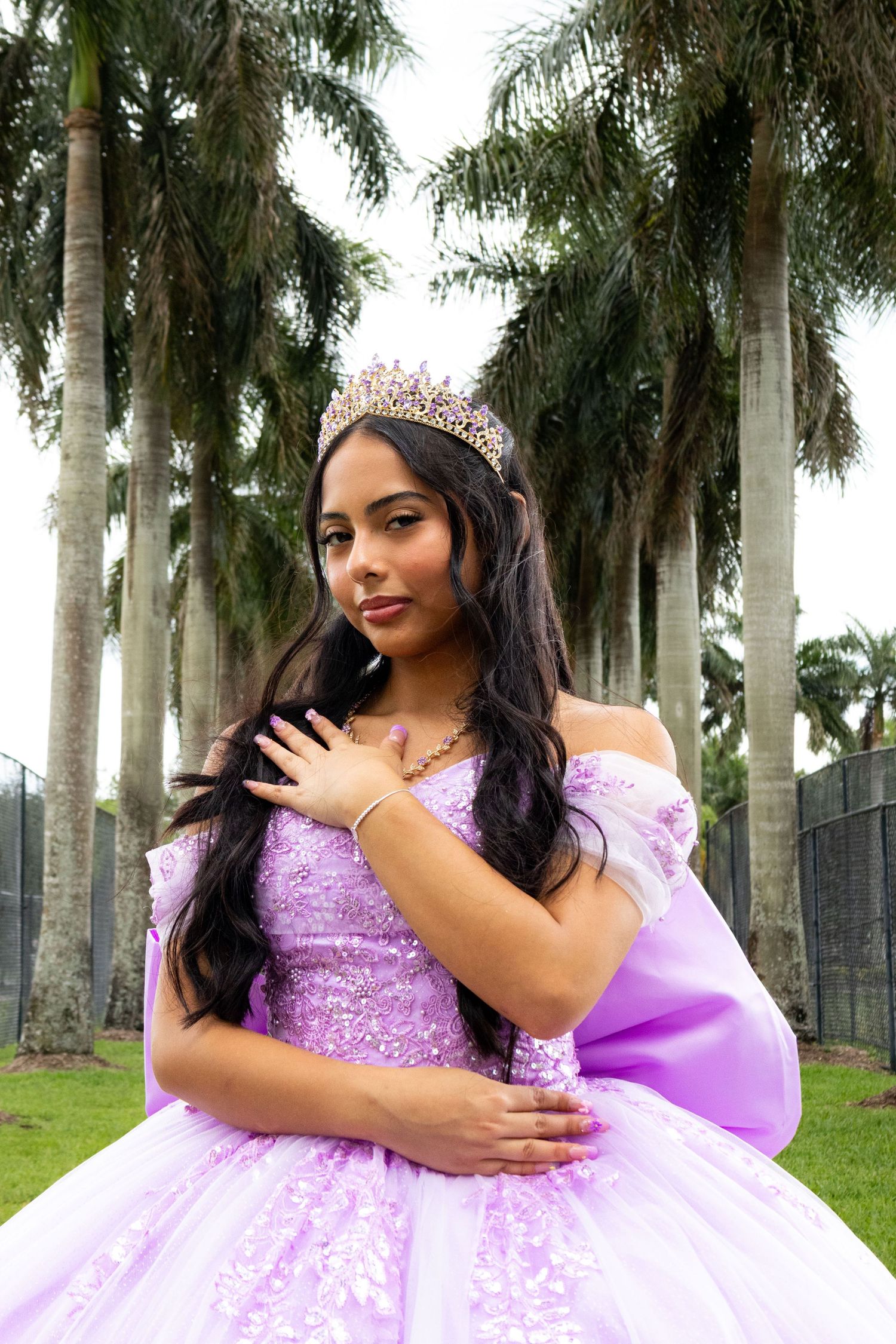 Young woman in lavender quinceañera ball gown with crown poses among tall palm trees outdoors.