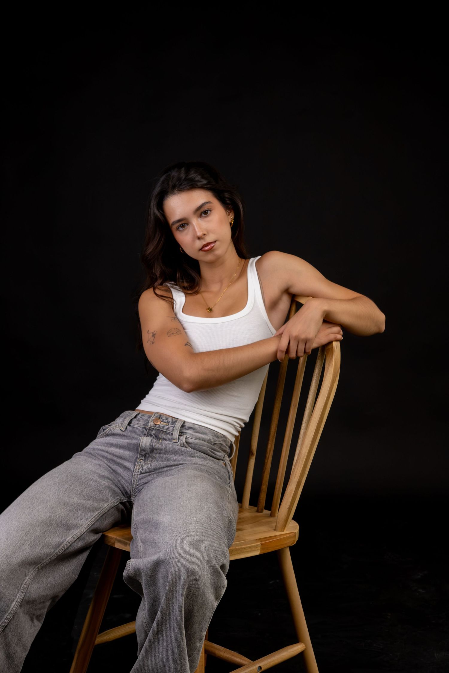 Mulher sentada numa cadeira, usando top branco e calças cinzentas, num fundo preto.