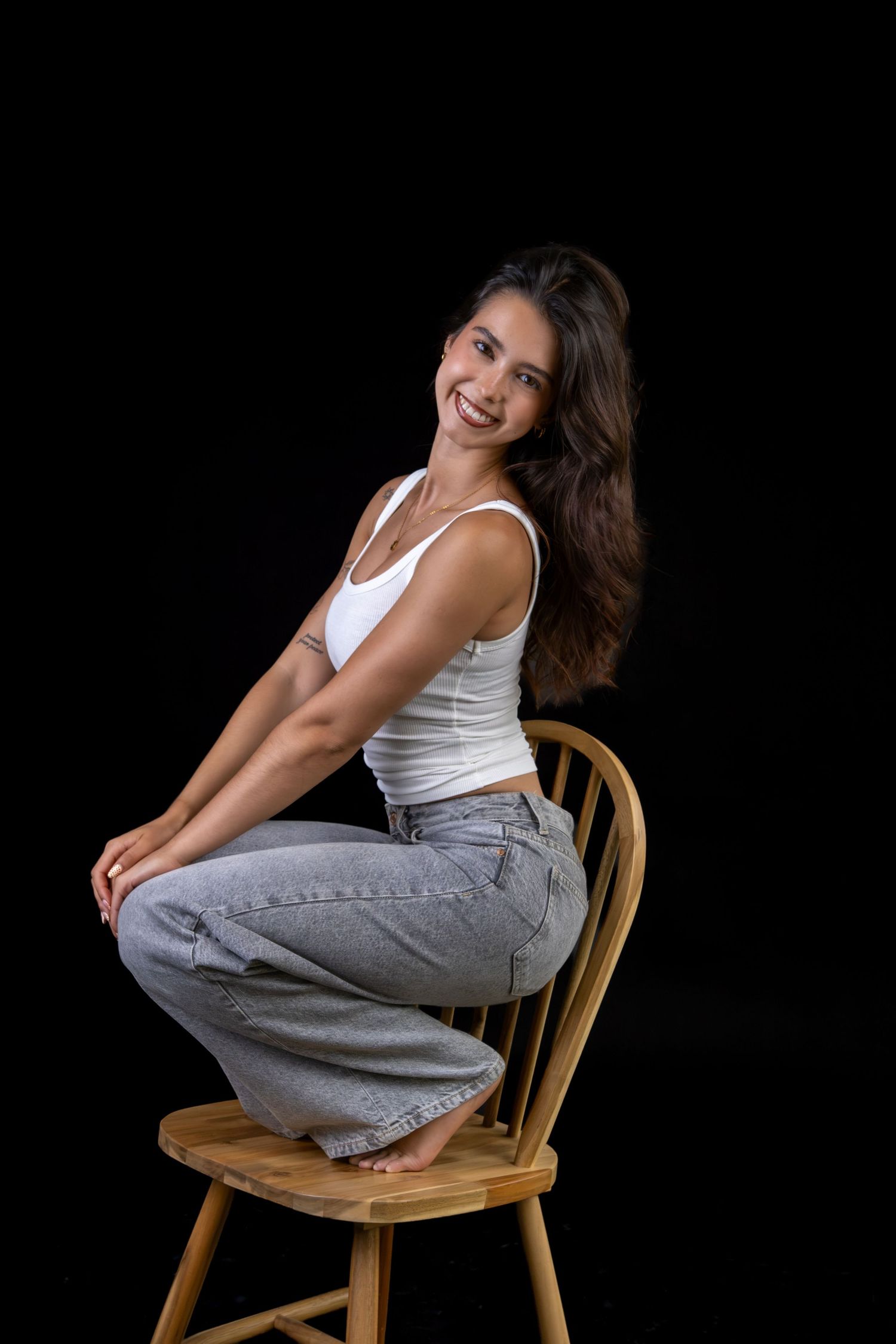 Mulher sentada numa cadeira de madeira, usando top branco e calças cinzentas.