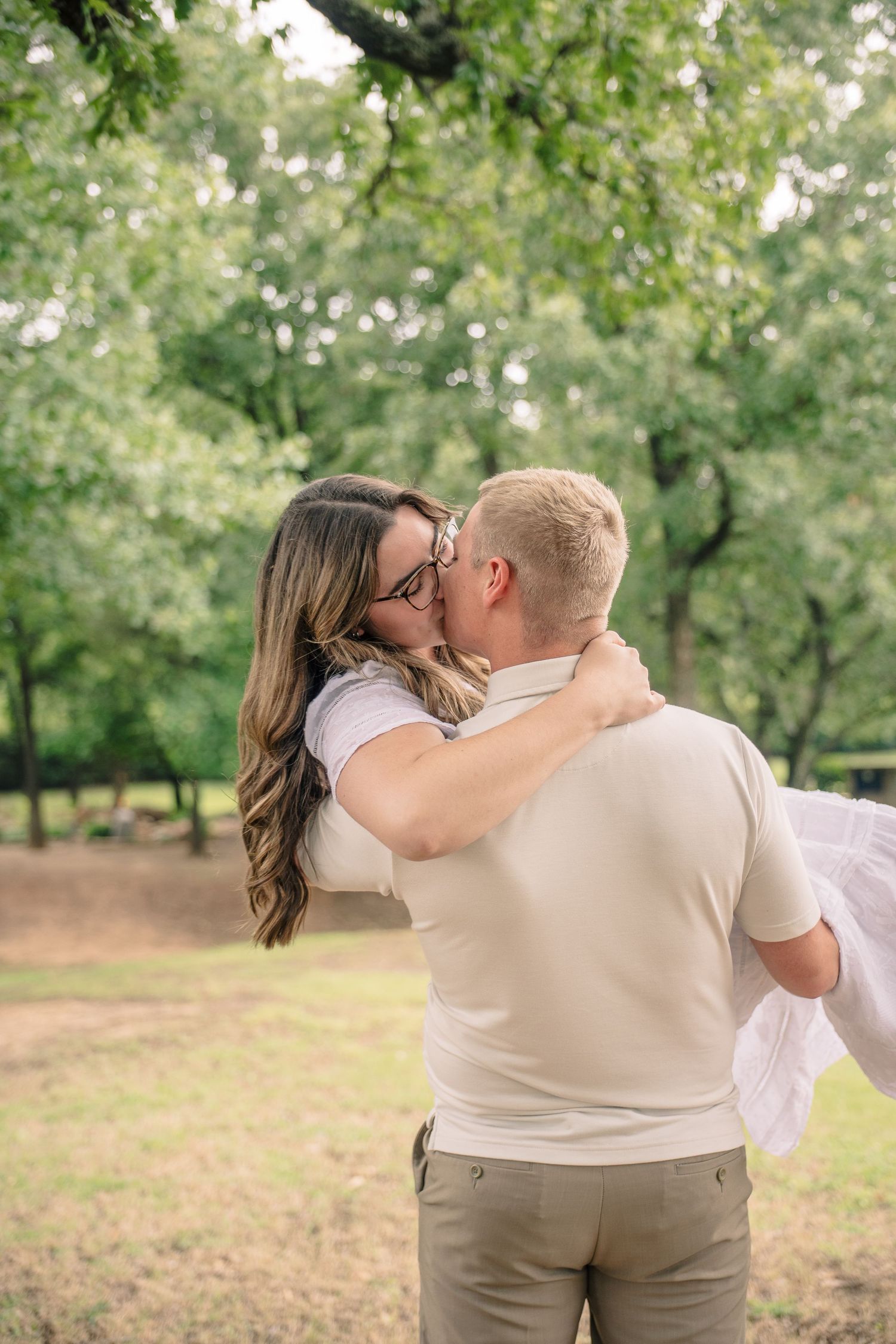 Man carrying woman in his arms while sharing a romantic kiss outdoors in a lush green park.