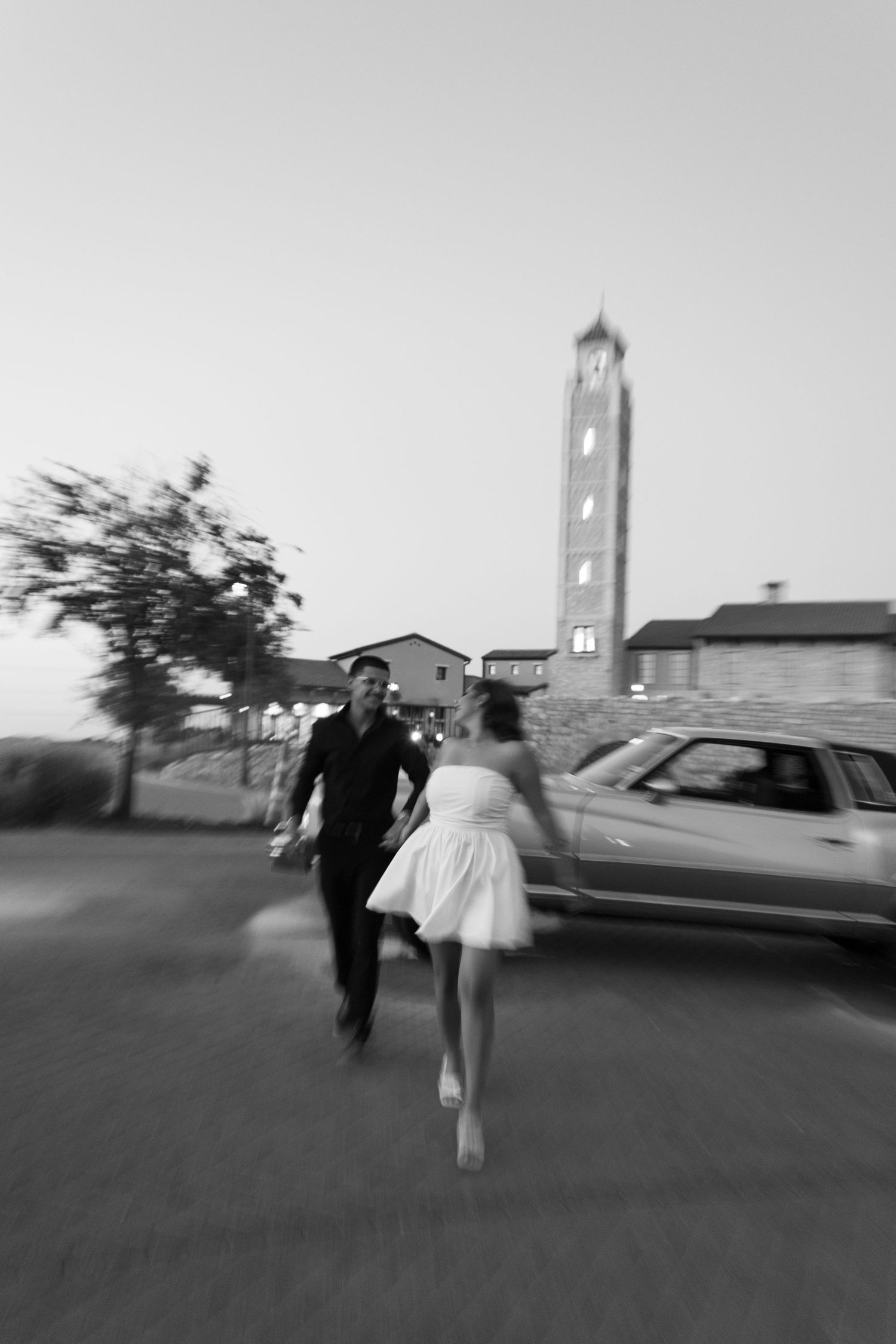 Couple running hand-in-hand near a vintage car with a tall clock tower in background.
