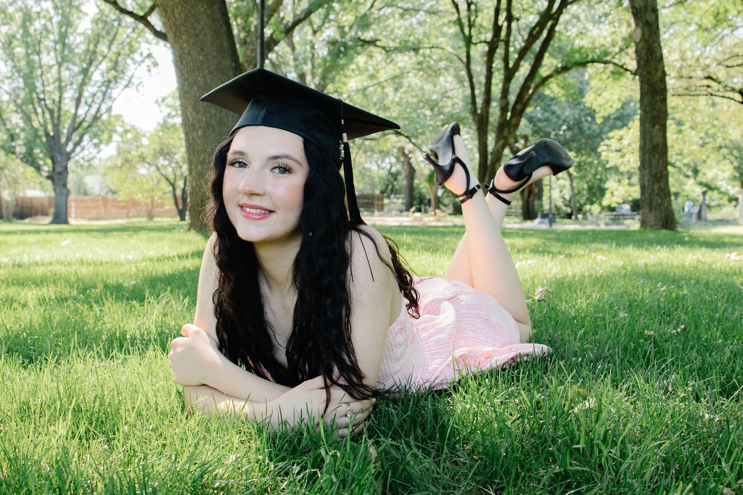 Graduate in cap and pink dress lying on green grass, smiling with legs raised behind her.