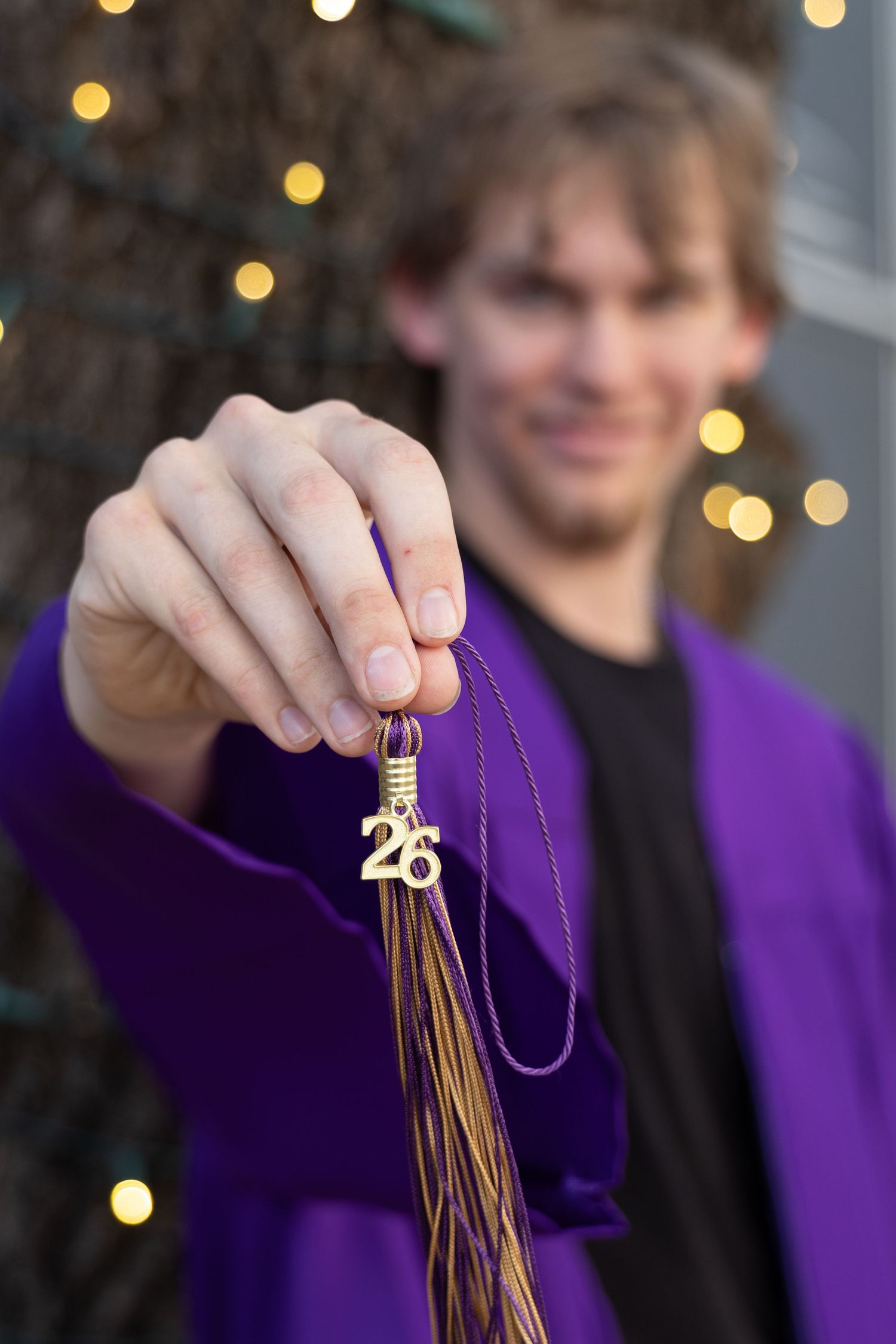 Close-up of graduate holding gold 26 tassel in purple gown with bokeh lights background.