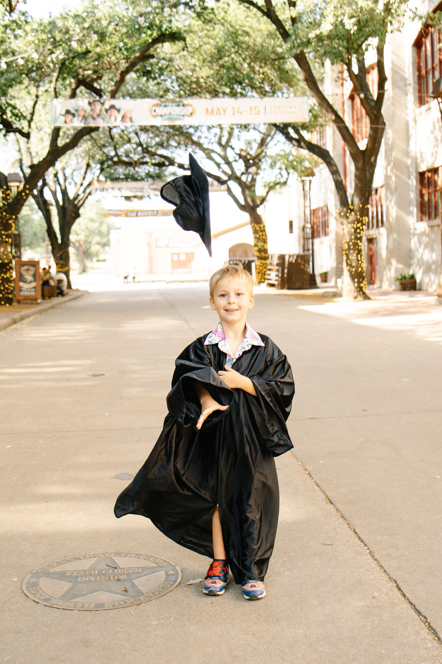 Young blonde boy in black graduation gown tossing cap on a tree-lined street.