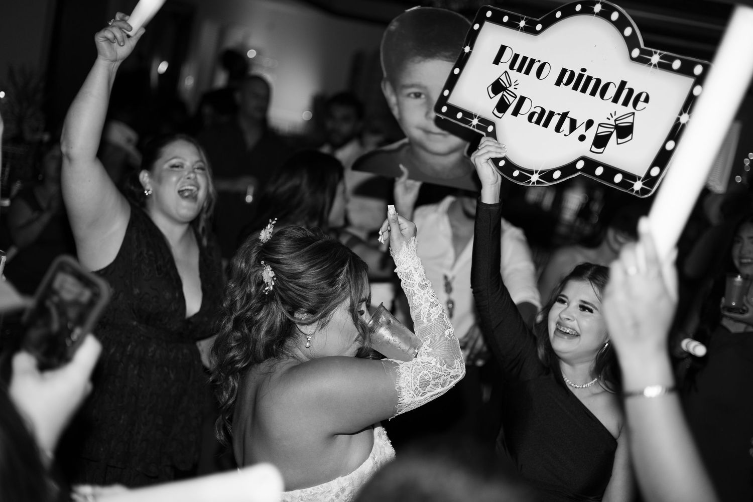 Bride celebrates on dance floor with guests holding a party sign at lively black and white wedding reception.