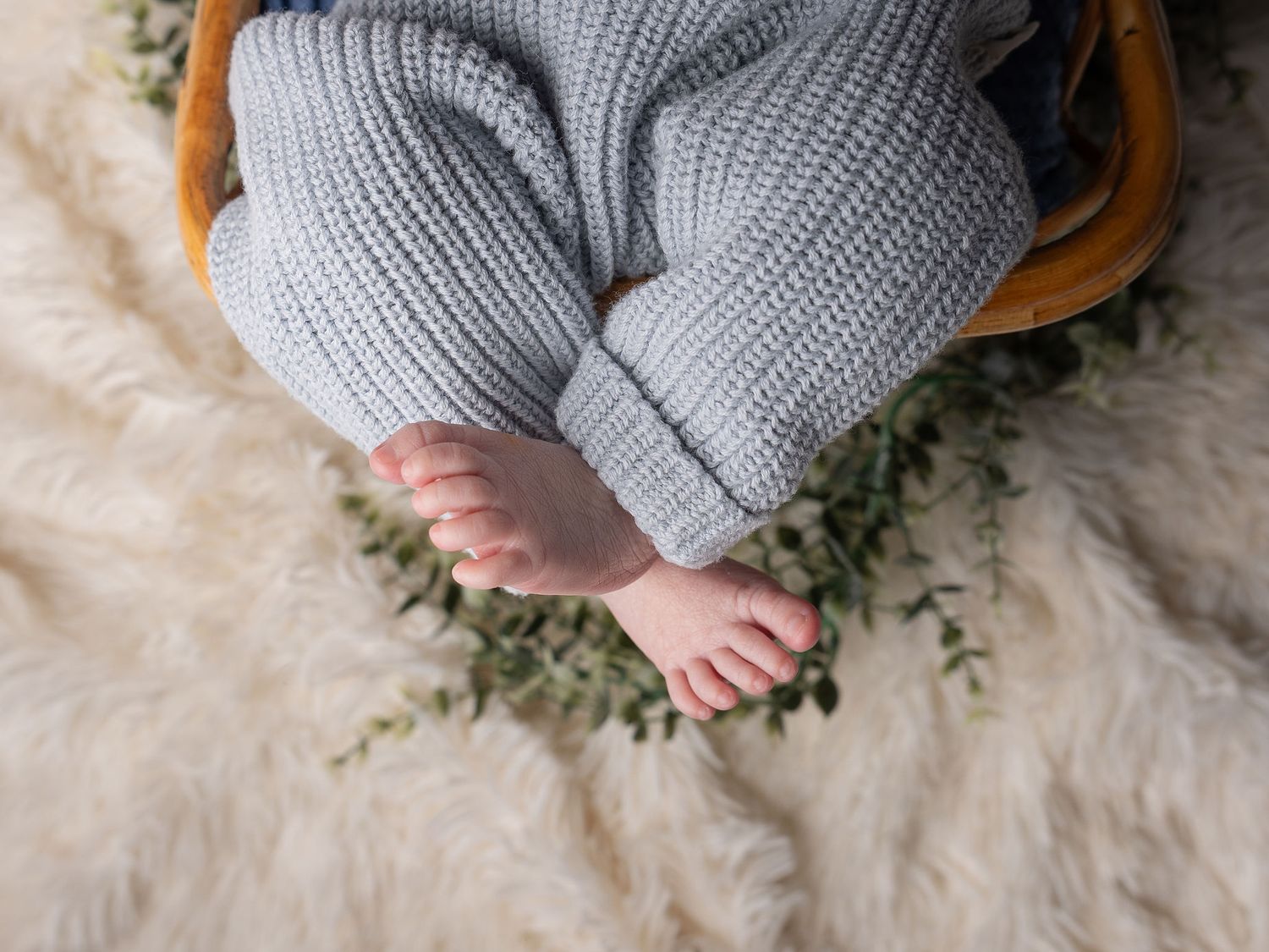 Newborn baby feet peeking out from gray knit pants in a wooden bowl, set on a cream faux fur rug with green eucalyptus leaves. Haslet Texas
