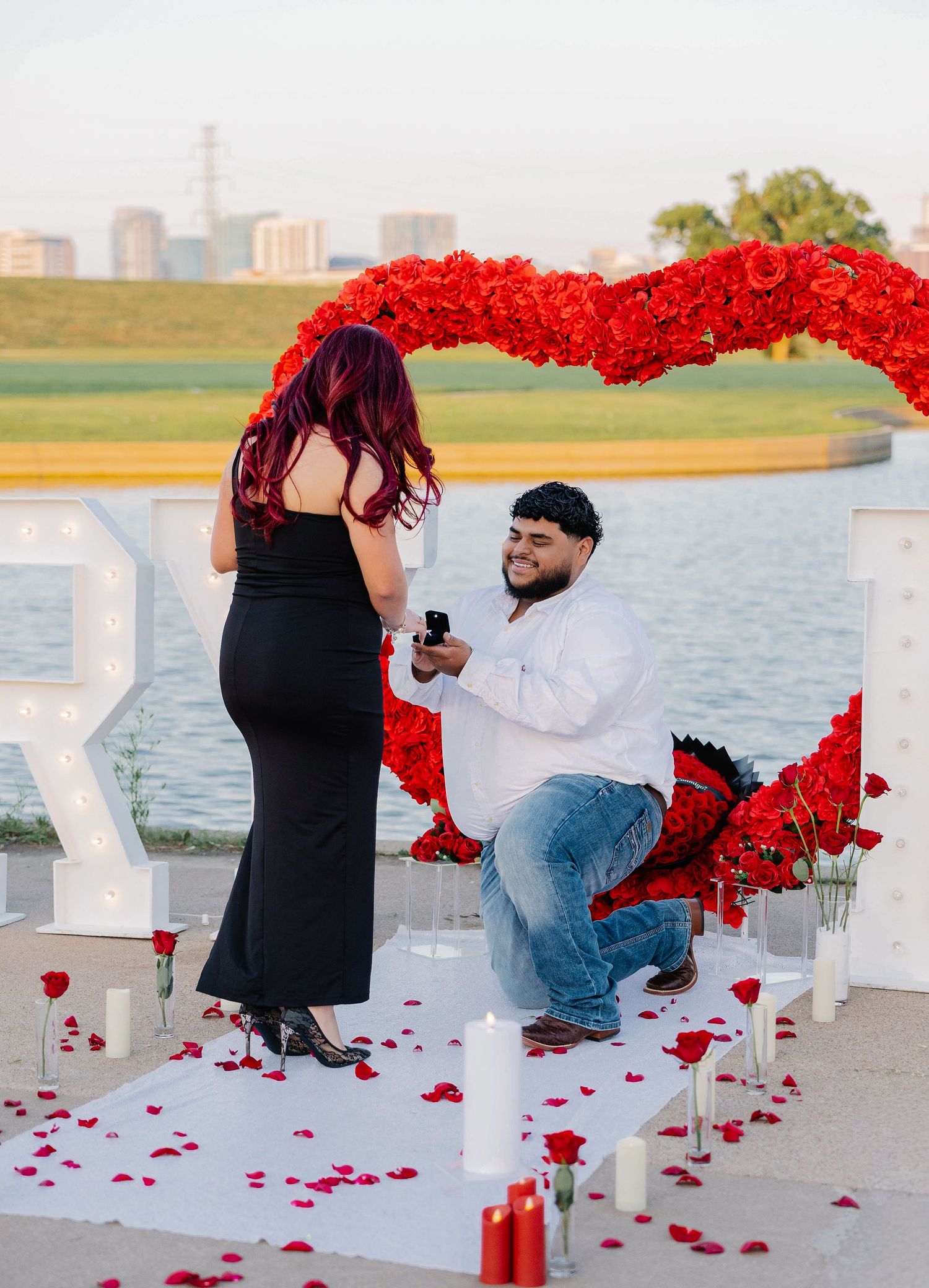 Man kneels with a ring box proposing to a woman beside a red rose heart arch, candles and petals by a lakeside at sunset. Dallas Texas