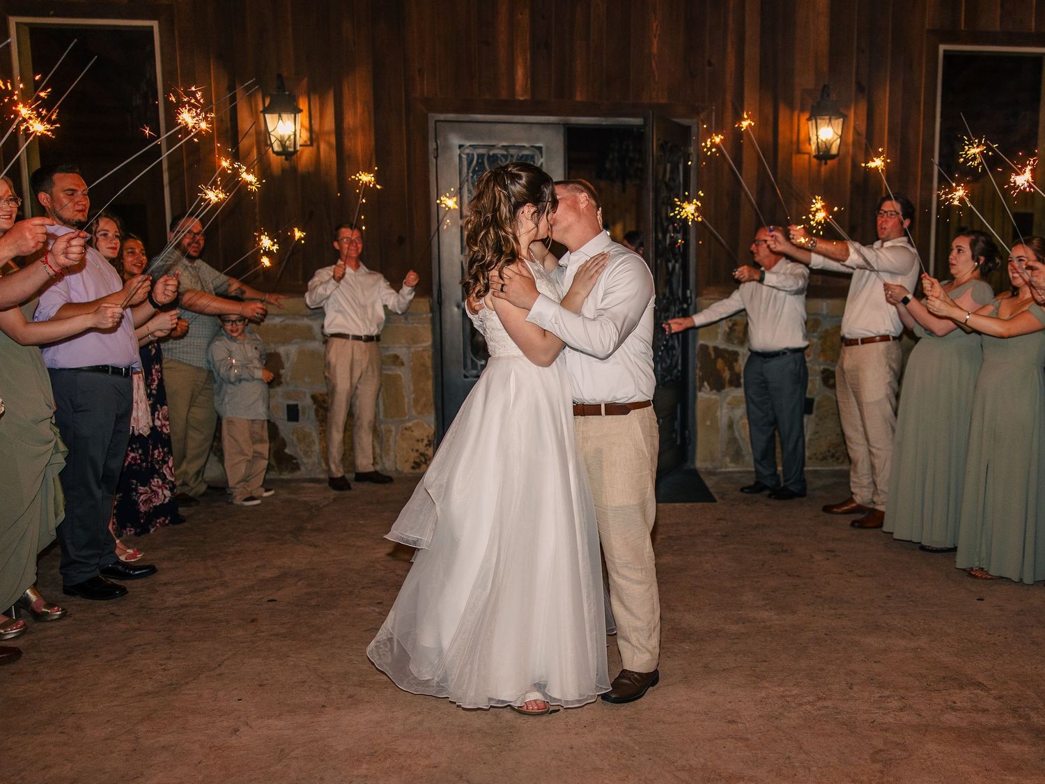 Bride and groom share a kiss on the dance floor as wedding guests hold lit sparklers in a warm, rustic wooden venue.