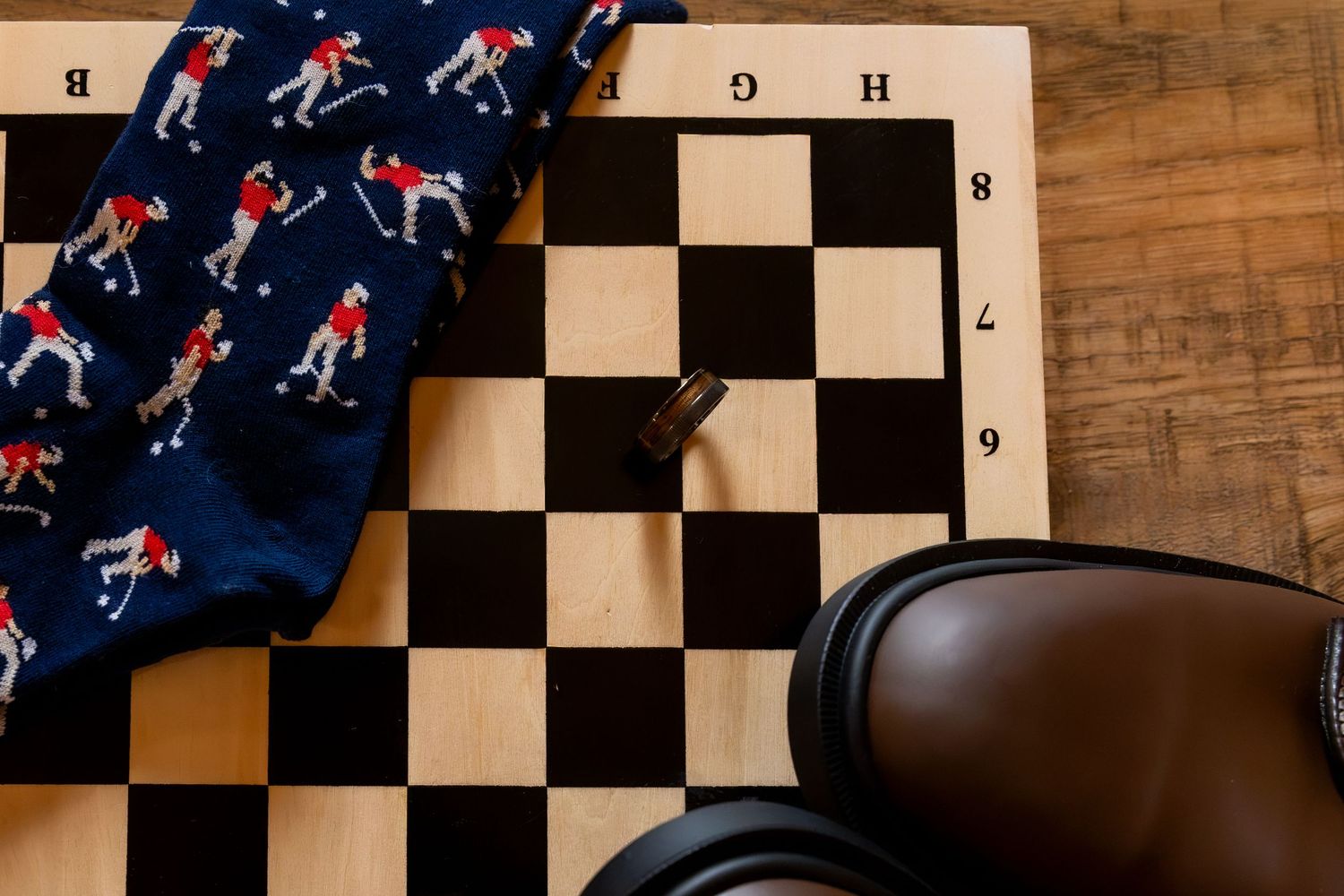 Navy socks with a red-shirted golfer pattern lie on a wooden chessboard beside a brown leather shoe and a black chess piece.