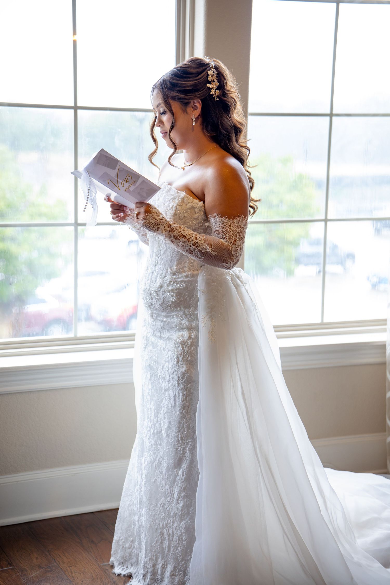 Bride in lace off-shoulder wedding gown reads a letter by a sunlit window on her wedding day.