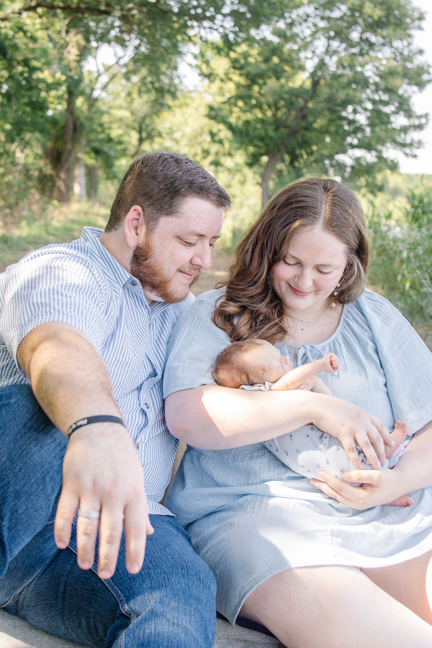 A couple sits outdoors cradling their newborn baby, smiling lovingly in a sunlit park setting.
