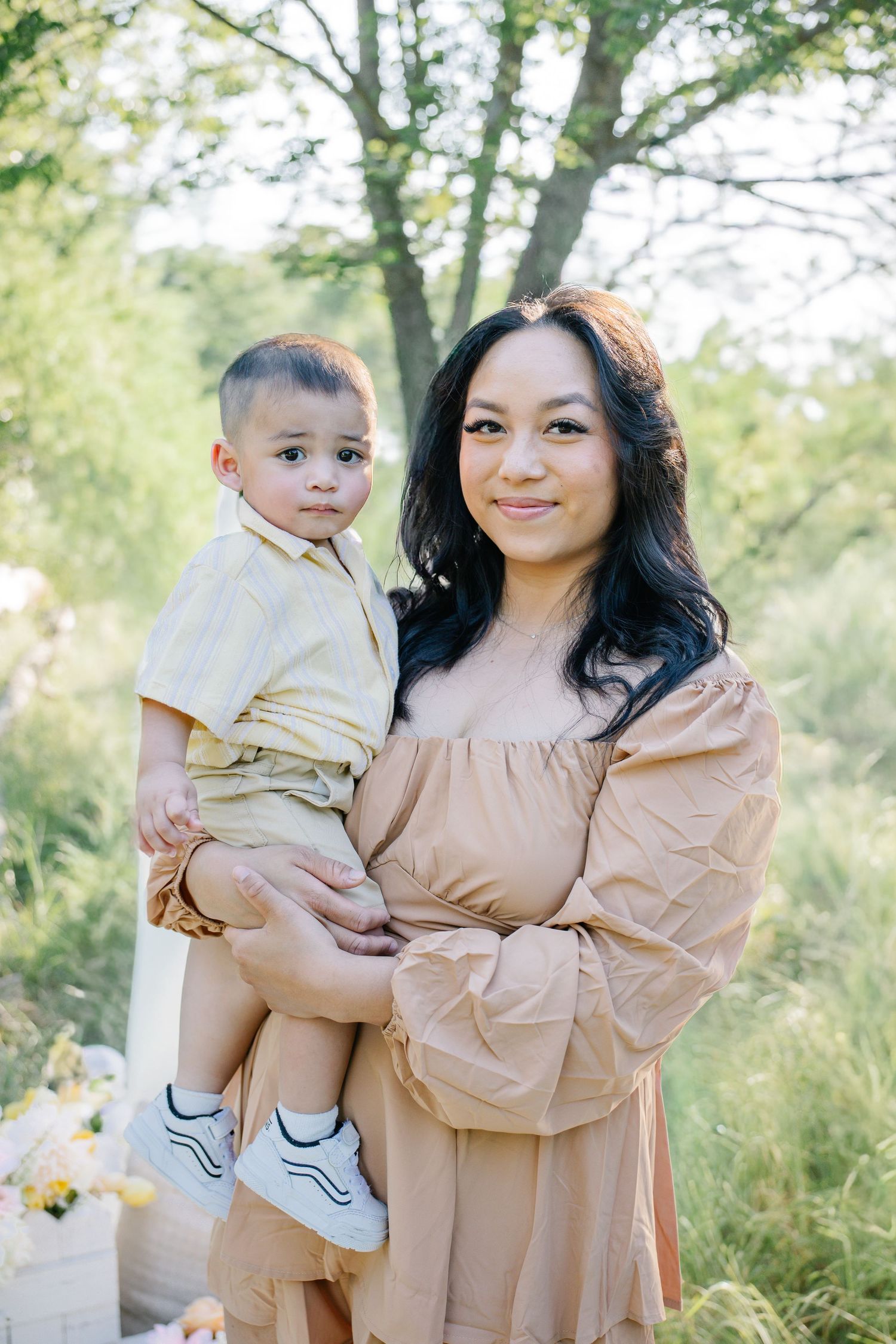 A woman in a tan dress holds a young boy in a beige outfit outdoors among green trees.