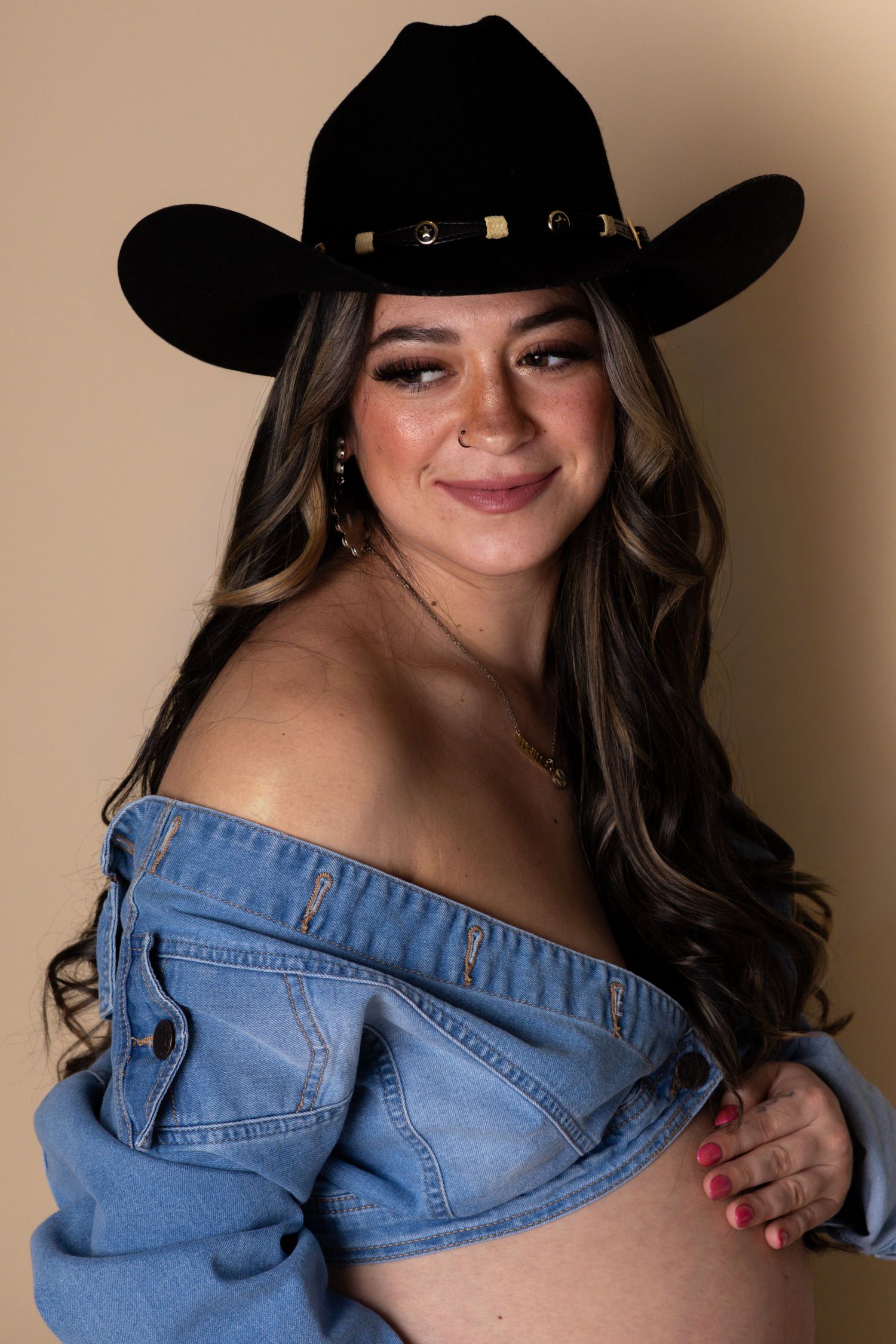 Smiling pregnant woman in black cowboy hat and off-shoulder denim top in studio portrait.