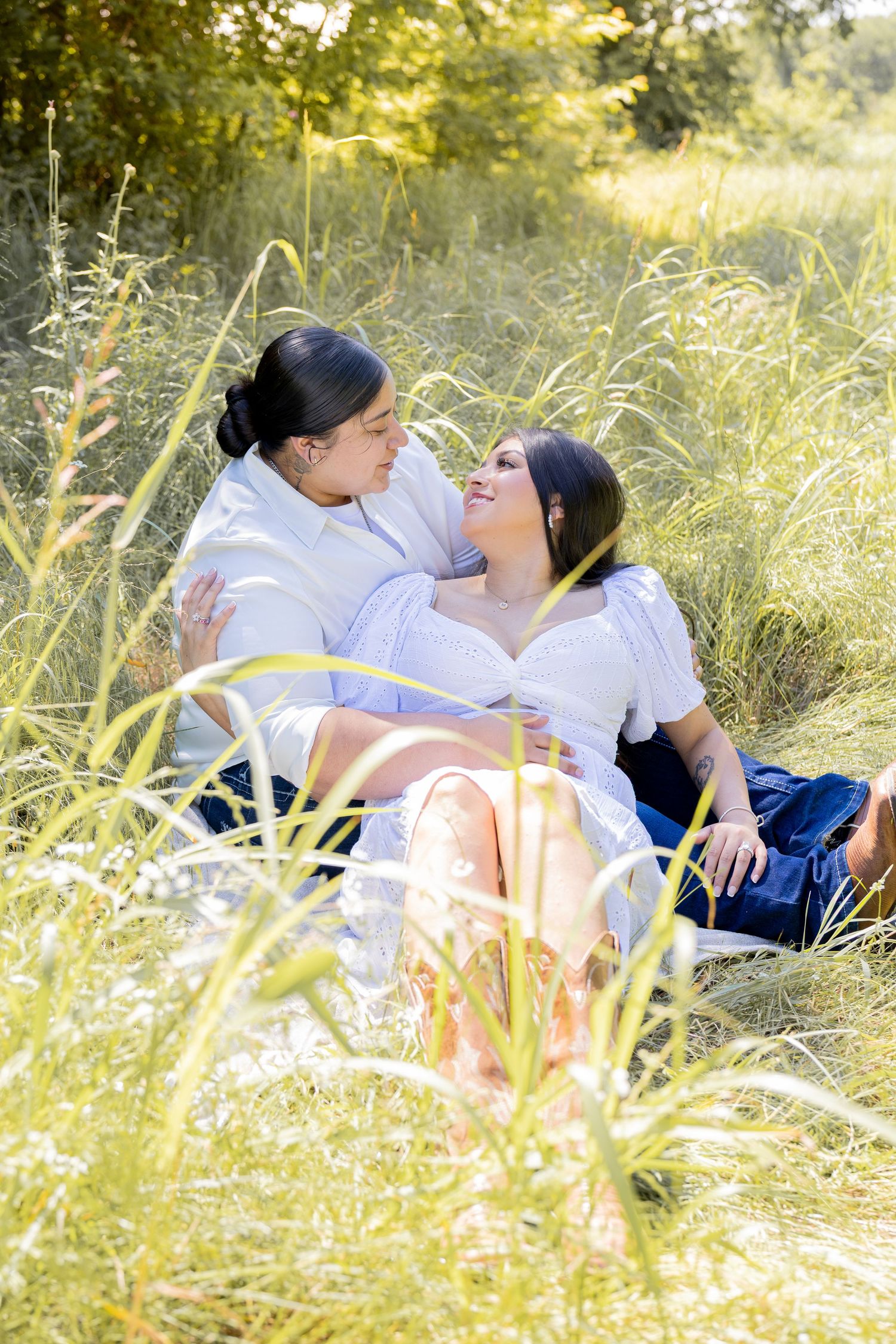 A couple sits closely together in a lush green meadow, gazing lovingly at each other outdoors.