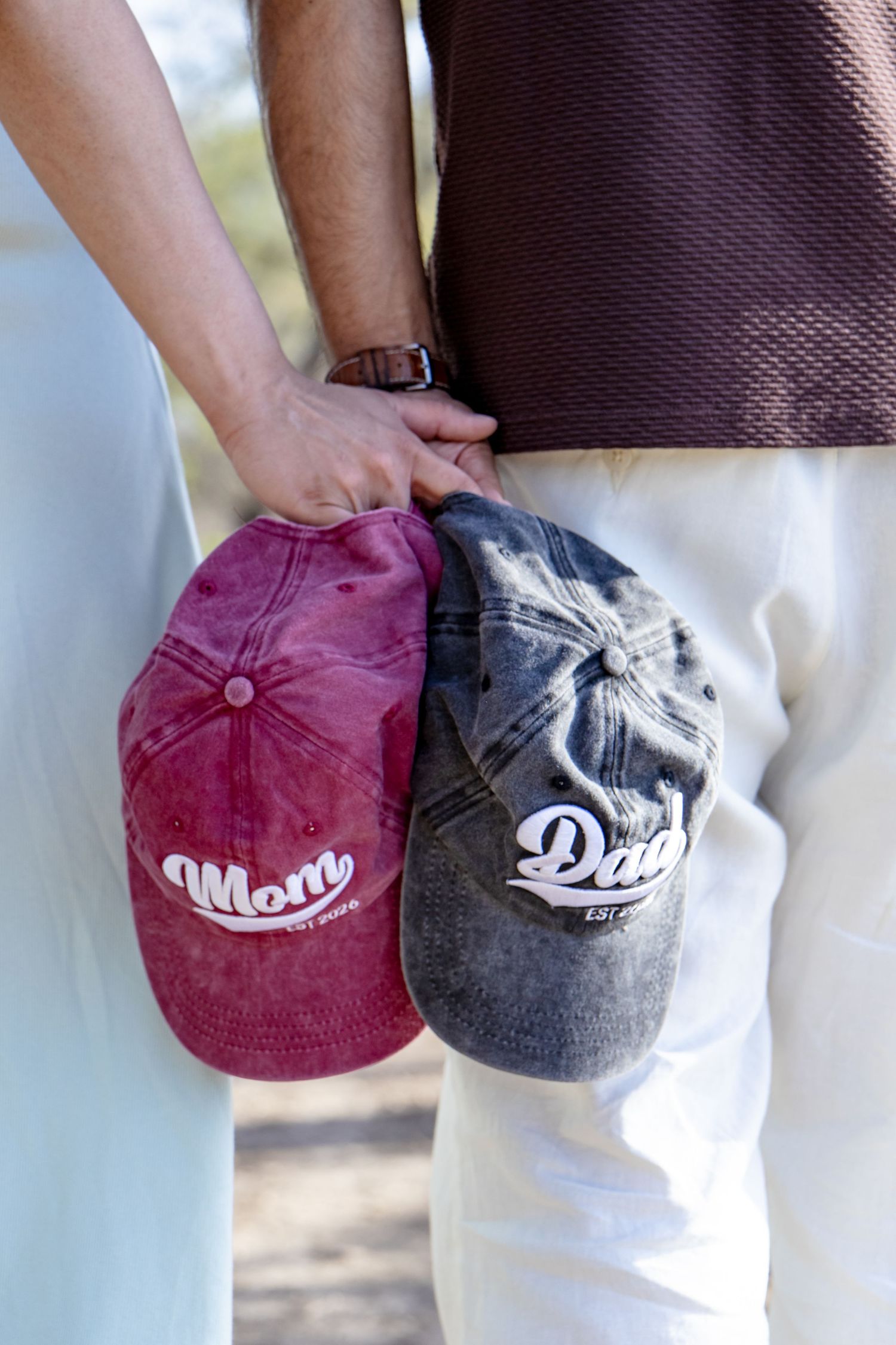 Close-up of expecting couple holding pink Mom and gray Dad baseball caps together at their sides.