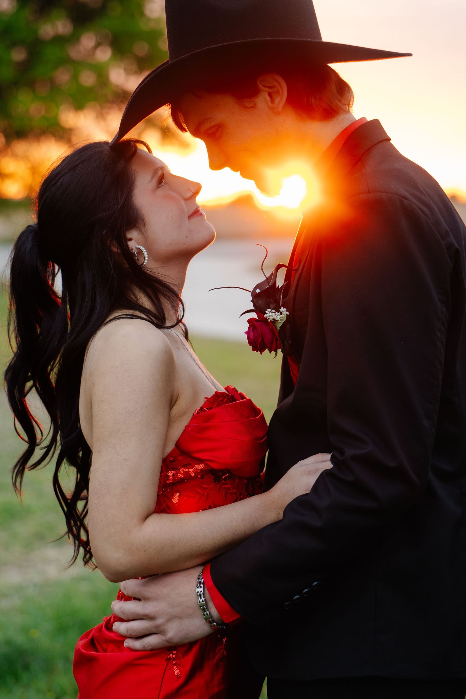 Romantic prom couple embracing at sunset, girl in red dress, boy in black cowboy hat with golden backlight.