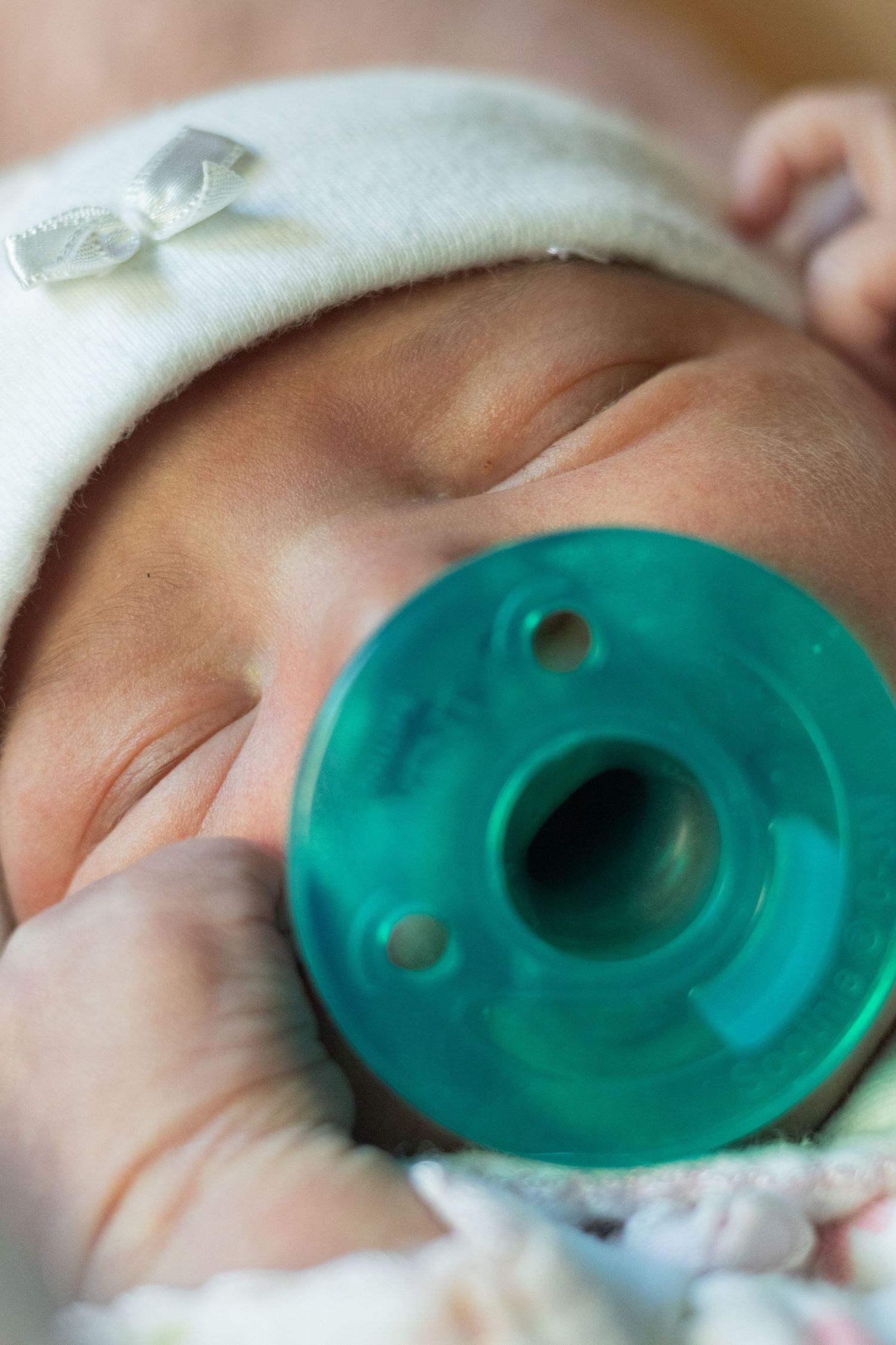 Newborn baby sleeping peacefully with a teal pacifier, wearing a white hat.