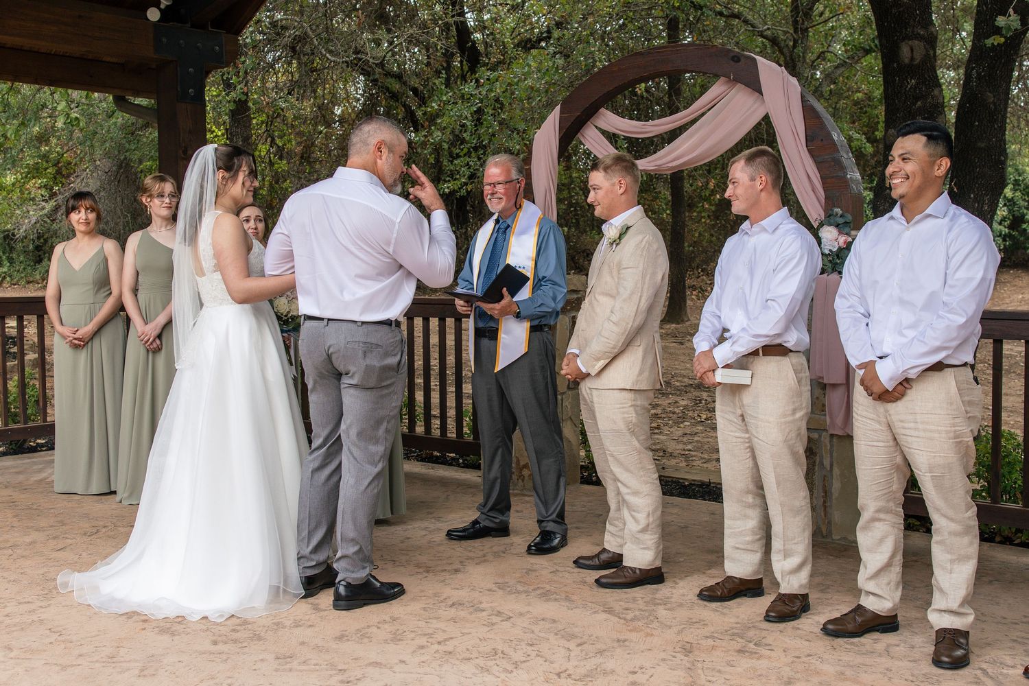 Bride in a white gown and groom exchange vows before an officiant at an outdoor wedding ceremony with bridesmaids and groomsmen beside a dra
