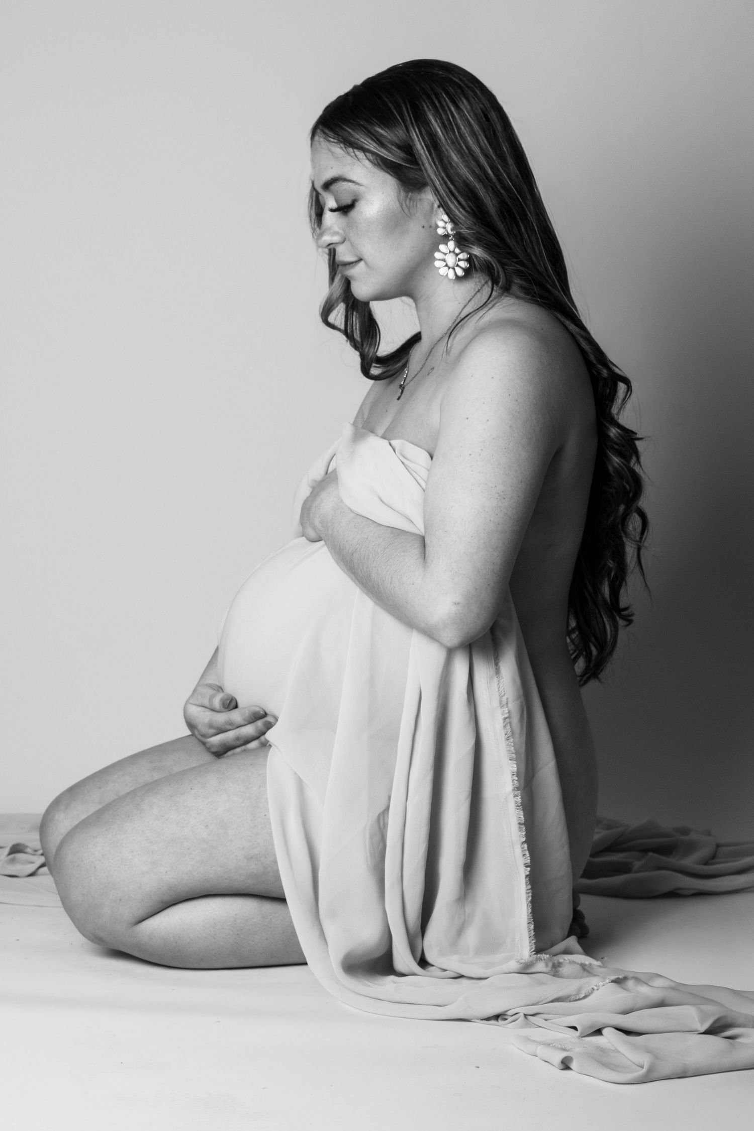 Black and white maternity photo of a pregnant woman kneeling, cradling her belly in a flowing dress.