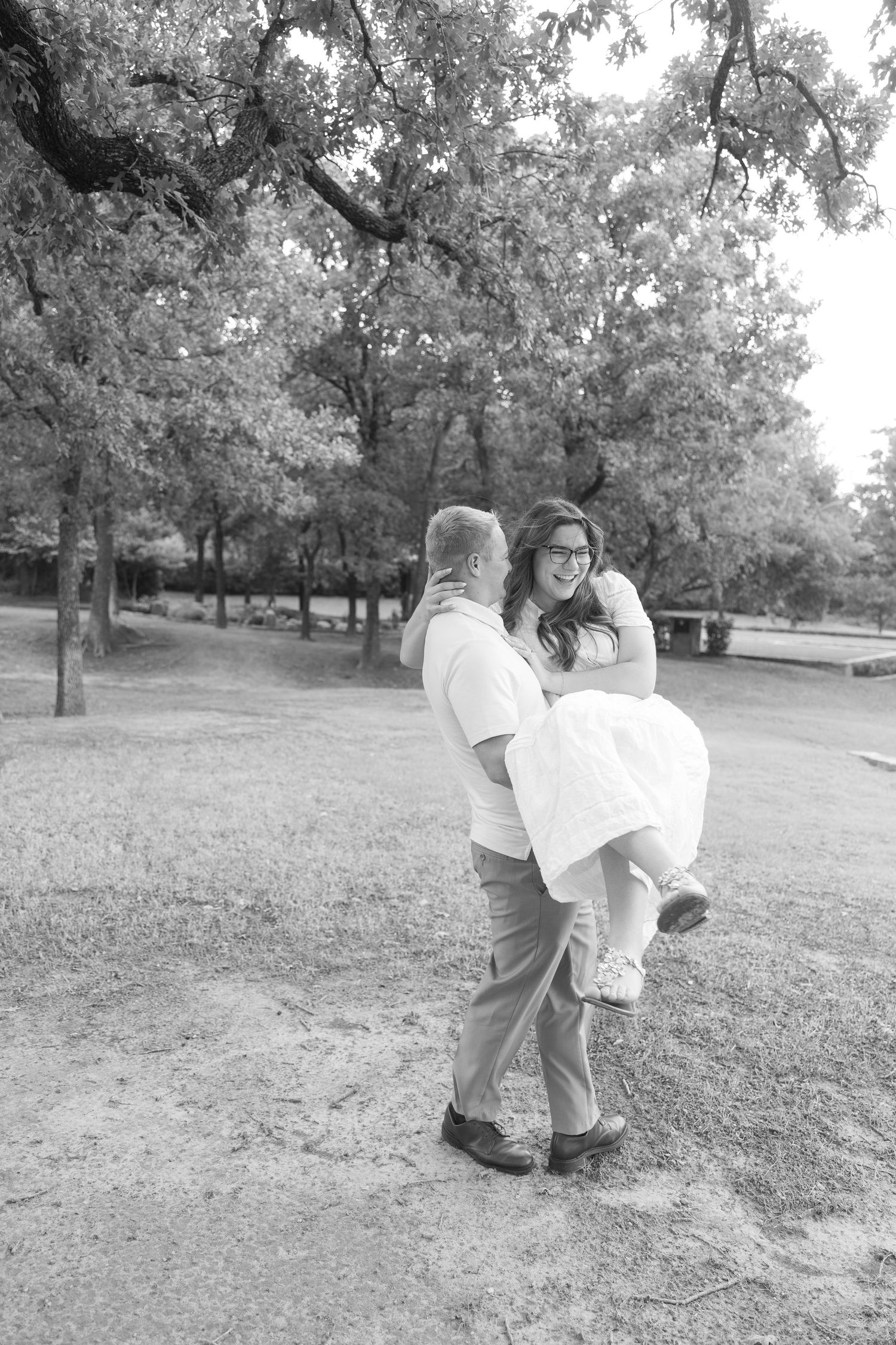 Couple sharing a joyful moment outdoors, man lifting woman in white dress, black and white photo in park setting.