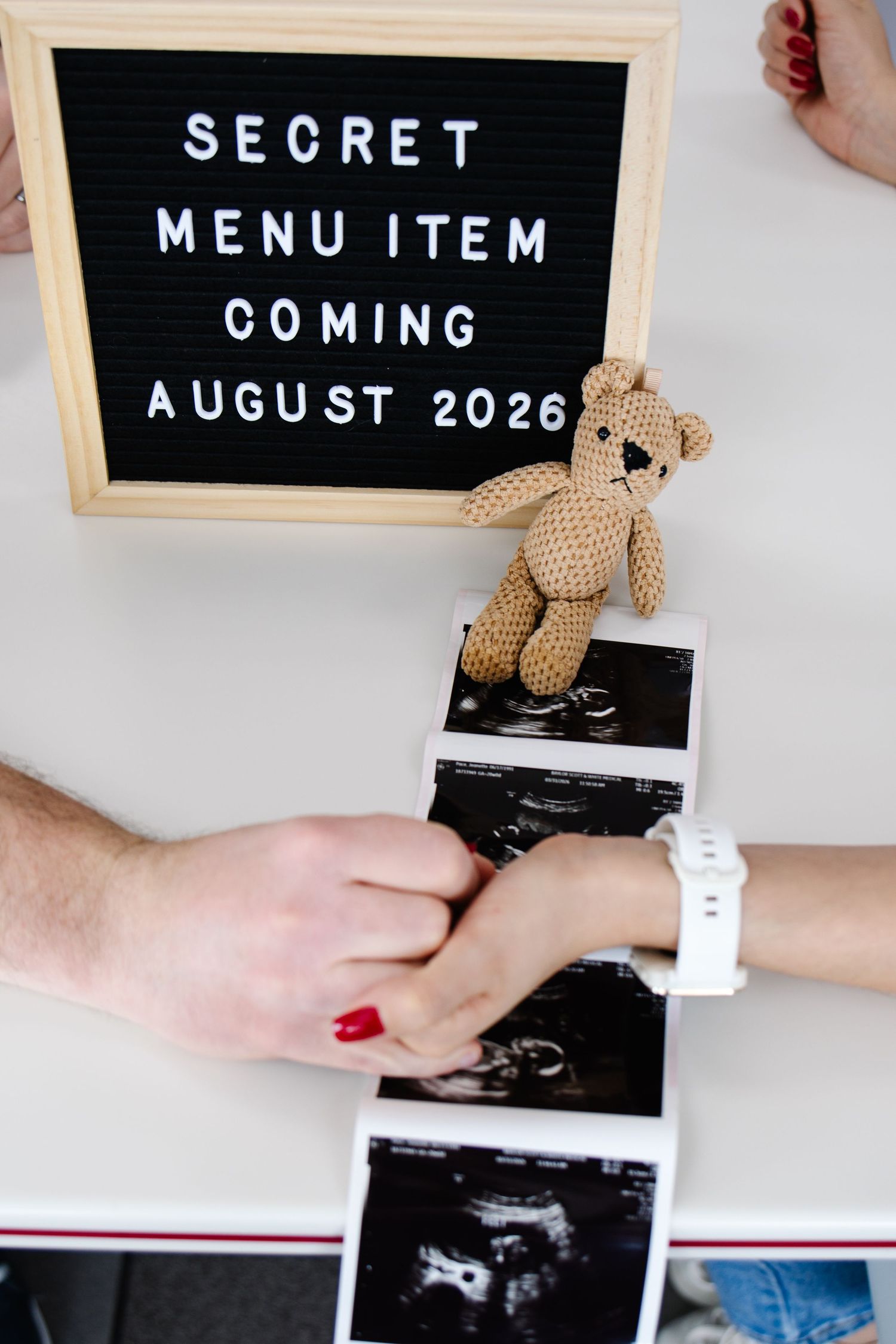 Couple holding hands over ultrasound photos beside a letter board announcing baby coming August 2026.