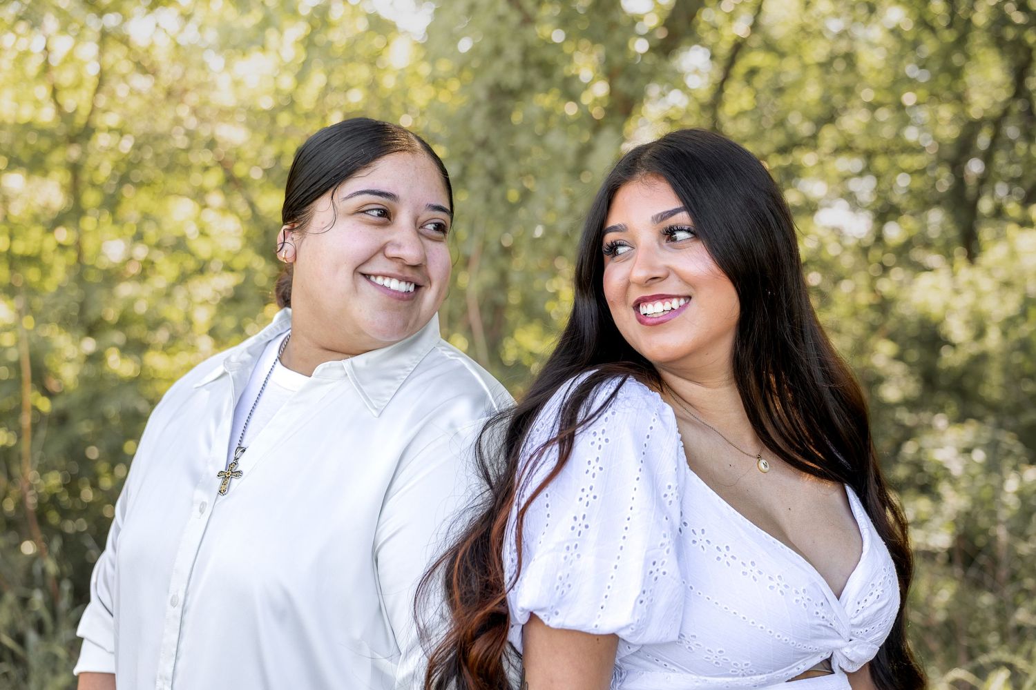 Two women smiling together outdoors surrounded by lush green trees in daylight.