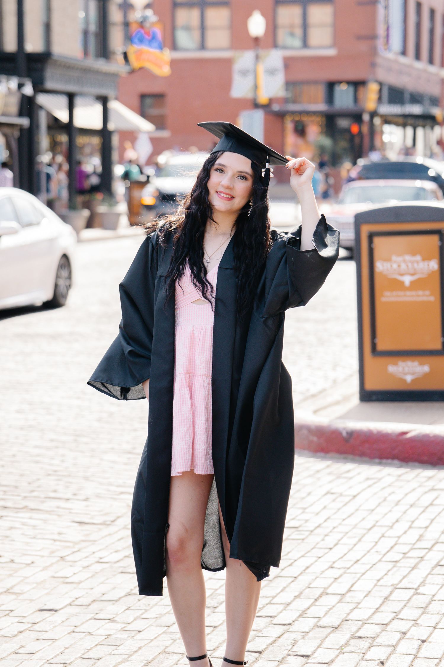 Graduate in black cap and gown over pink dress posing on brick street in urban setting.
