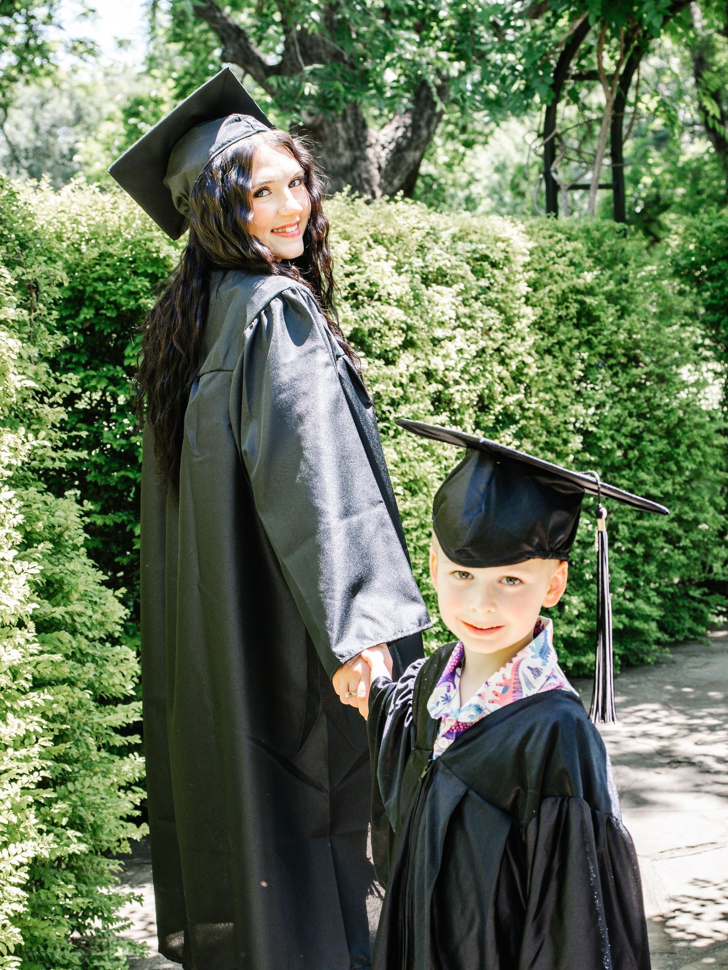 Woman and young boy both wearing black graduation caps and gowns posing together outdoors.