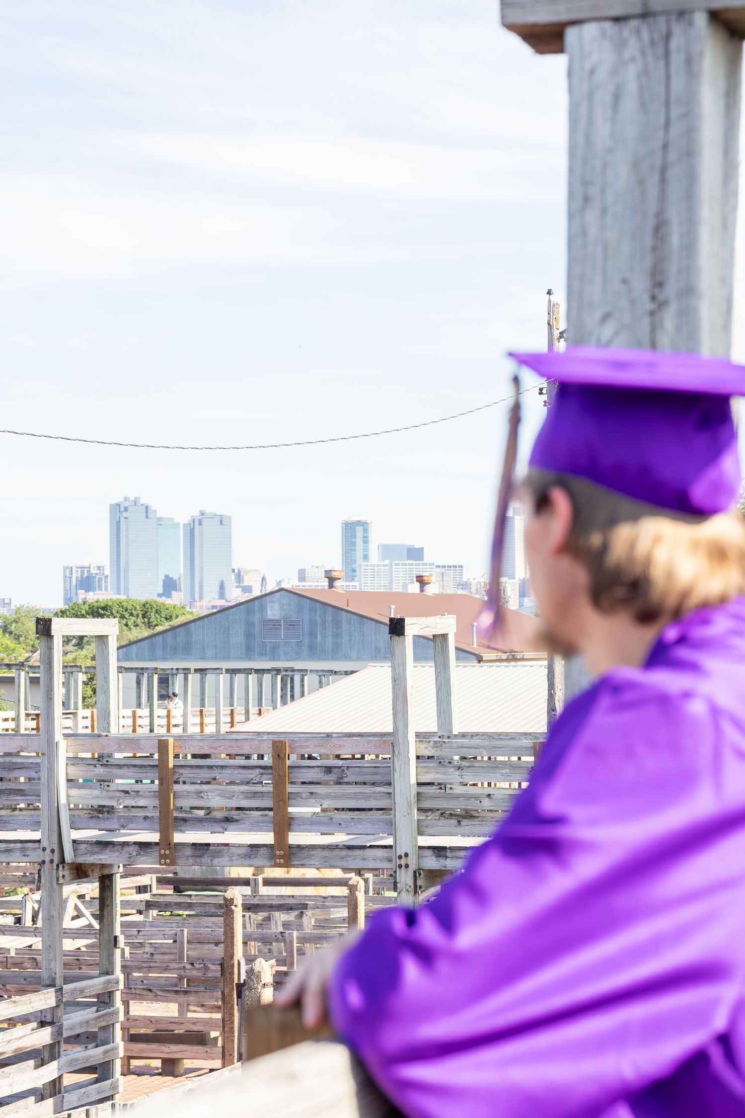 Graduate in purple cap and gown overlooking city skyline from wooden overlook structure.