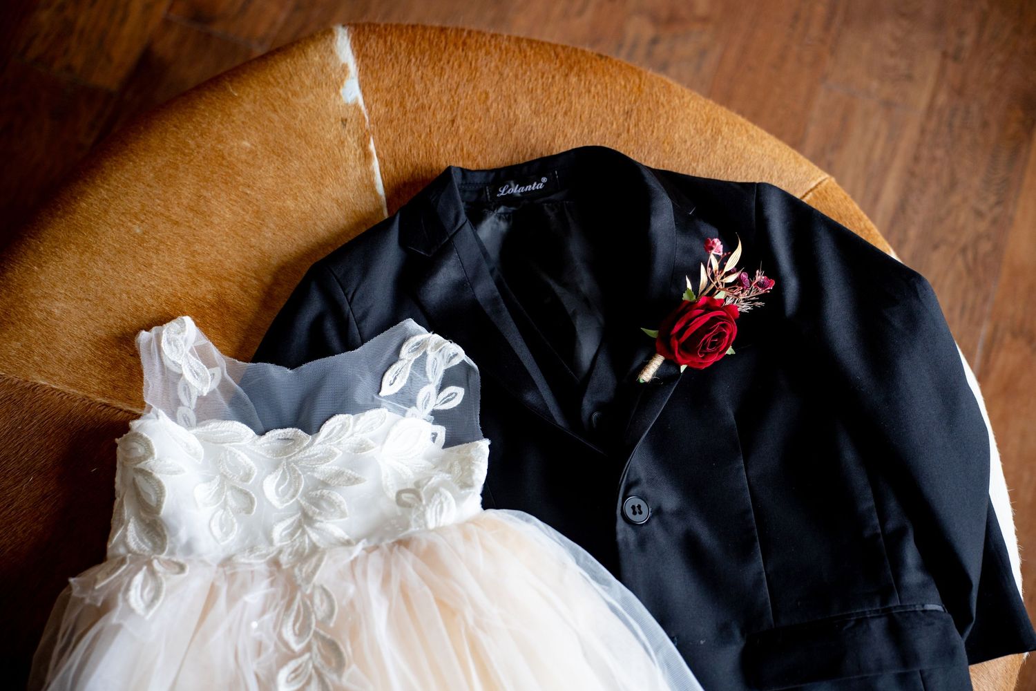 A white flower girl dress and black tuxedo jacket with red boutonniere displayed on a wooden chair for a wedding.
