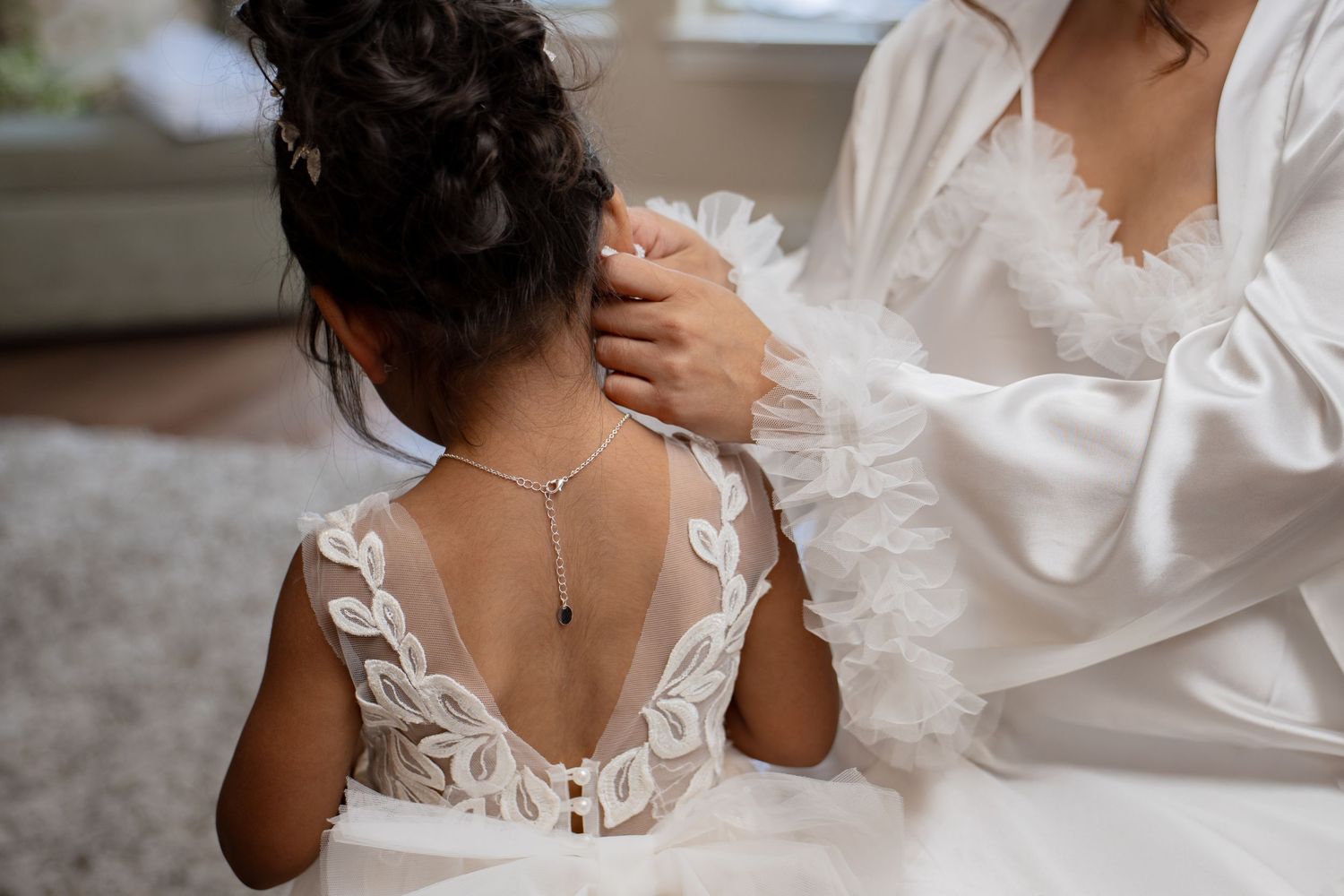 Woman in white robe fastening a necklace on a young girl in a white lace flower girl dress.