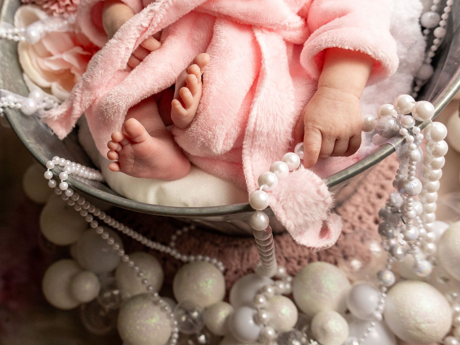 Newborn baby in pink outfit sleeping in a metal bowl surrounded by white pearl necklaces and decorative balls.