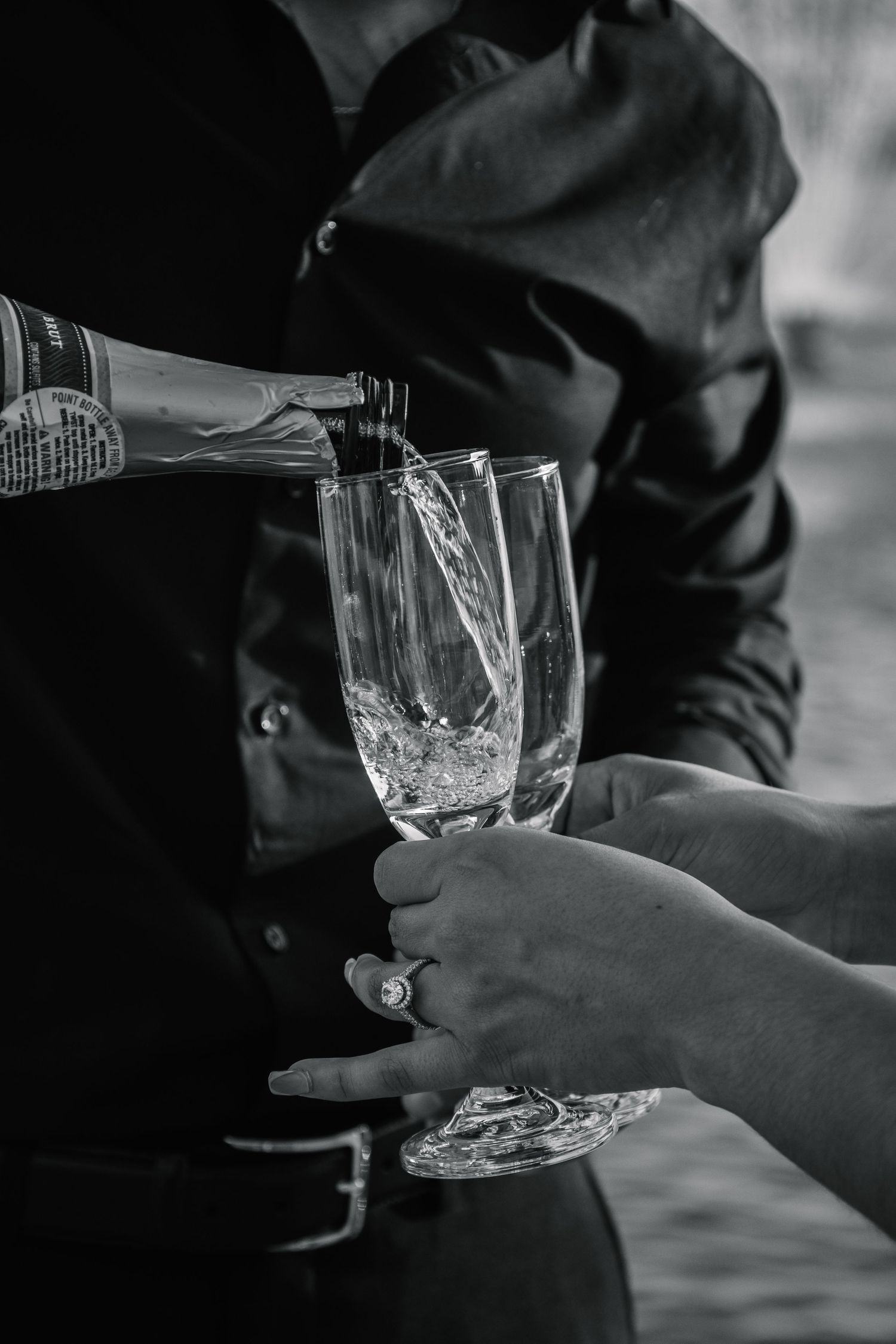 Black and white photo of champagne being poured into crystal flutes held by hands wearing an engagement ring.