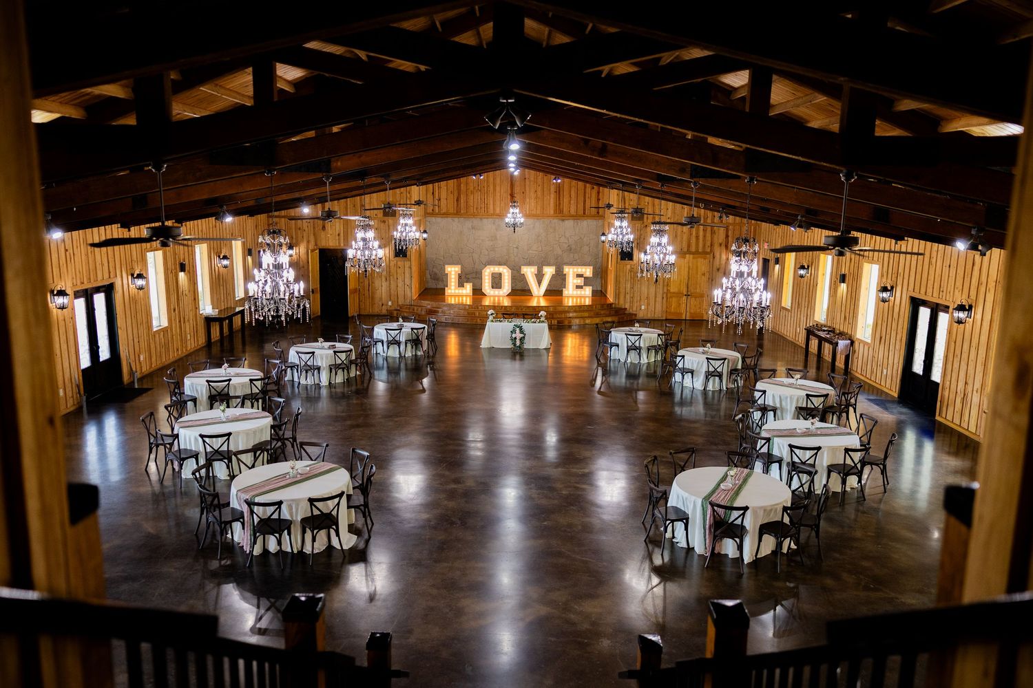 Rustic wooden wedding venue with round tables, chandeliers, and a lit LOVE sign ready for a reception.