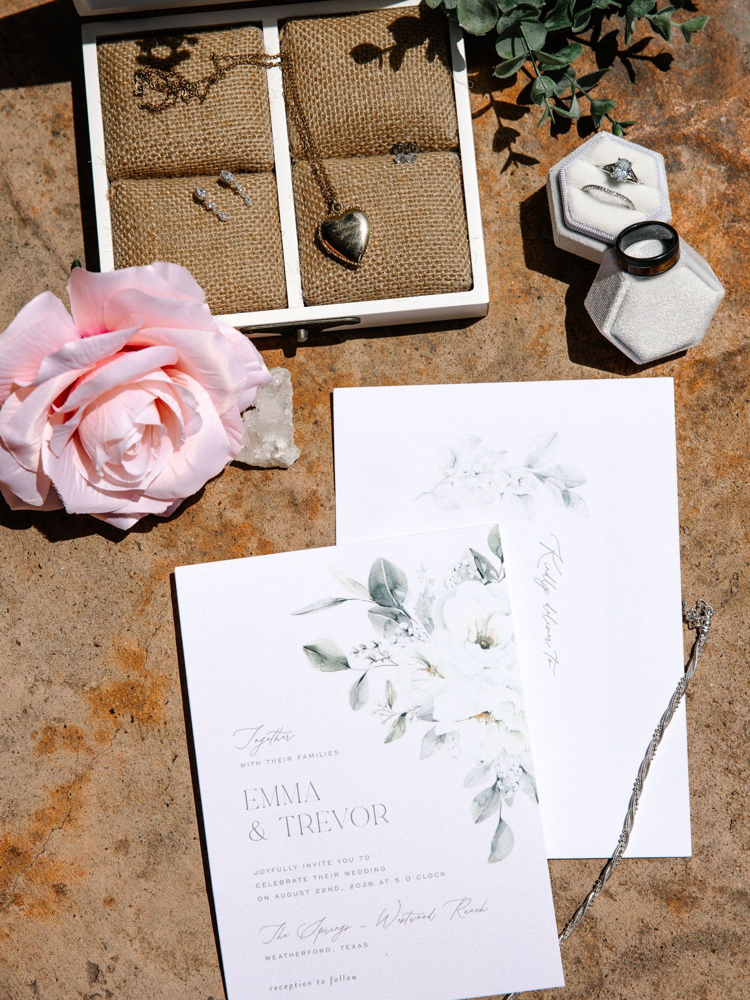 Elegant wedding stationery flat lay with Emma and Trevor's invitation, a pink rose, jewelry display tray and ring boxes.