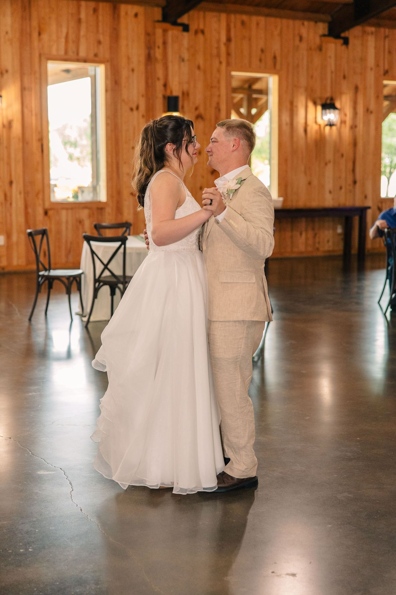 A bride in a white gown and a groom in a tan suit share a slow dance in a rustic wood-paneled venue.