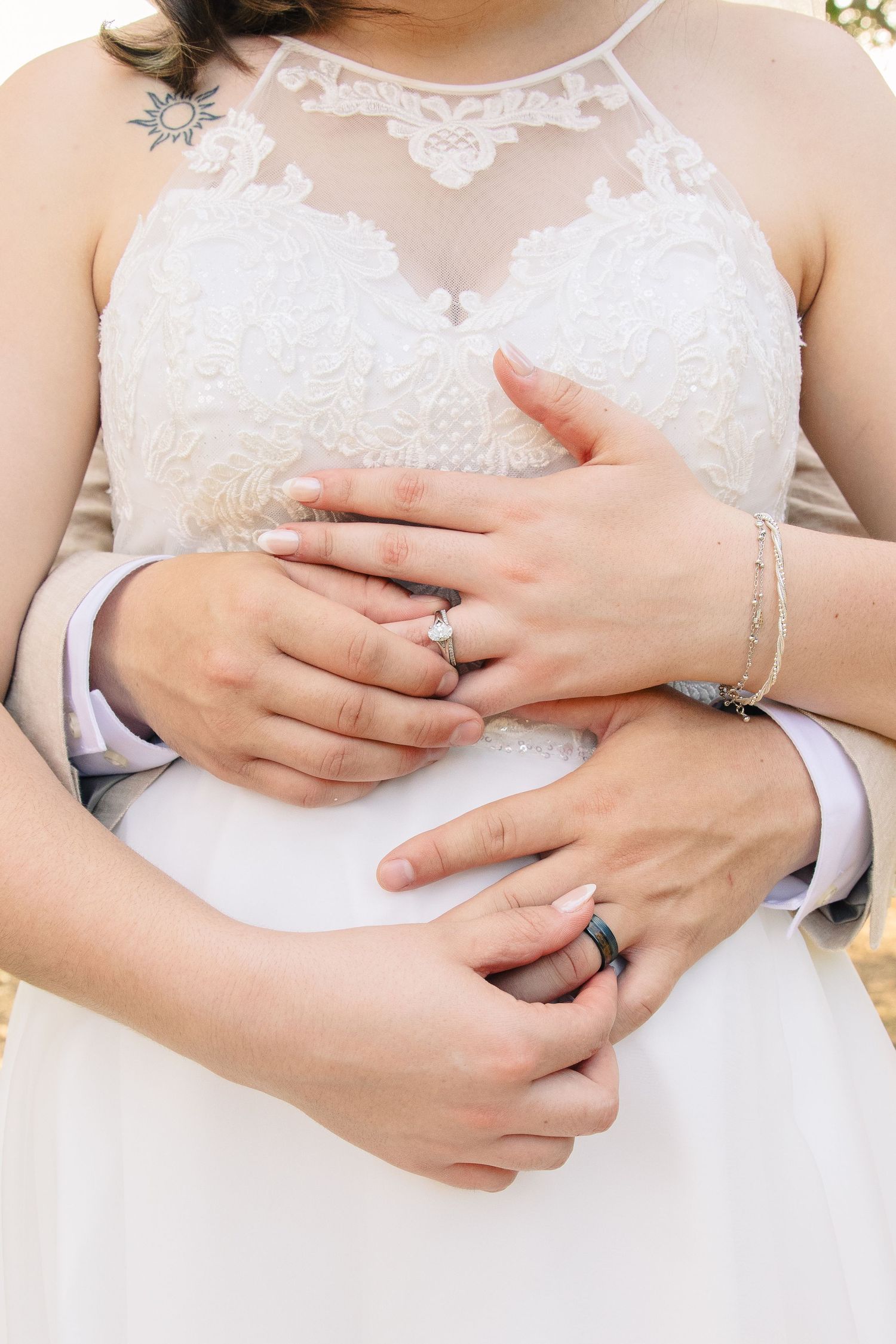 Bride in a white lace halter wedding dress with hands clasped over her waist as her partner embraces her, showing wedding rings.