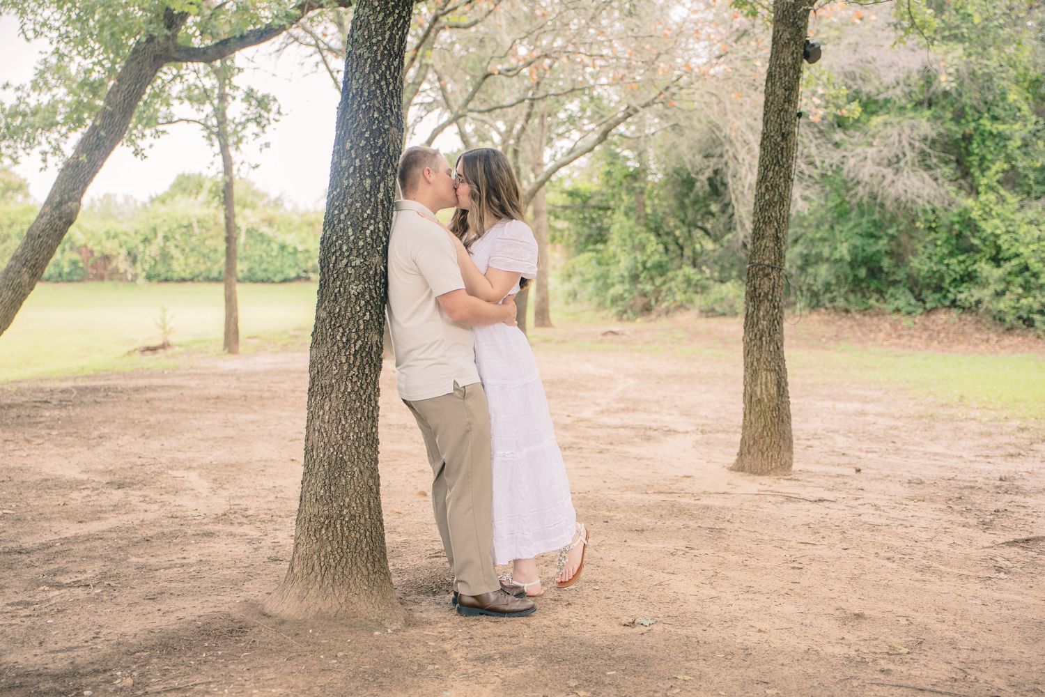 Couple sharing a romantic kiss leaning against a tree trunk in a sunlit outdoor park setting.