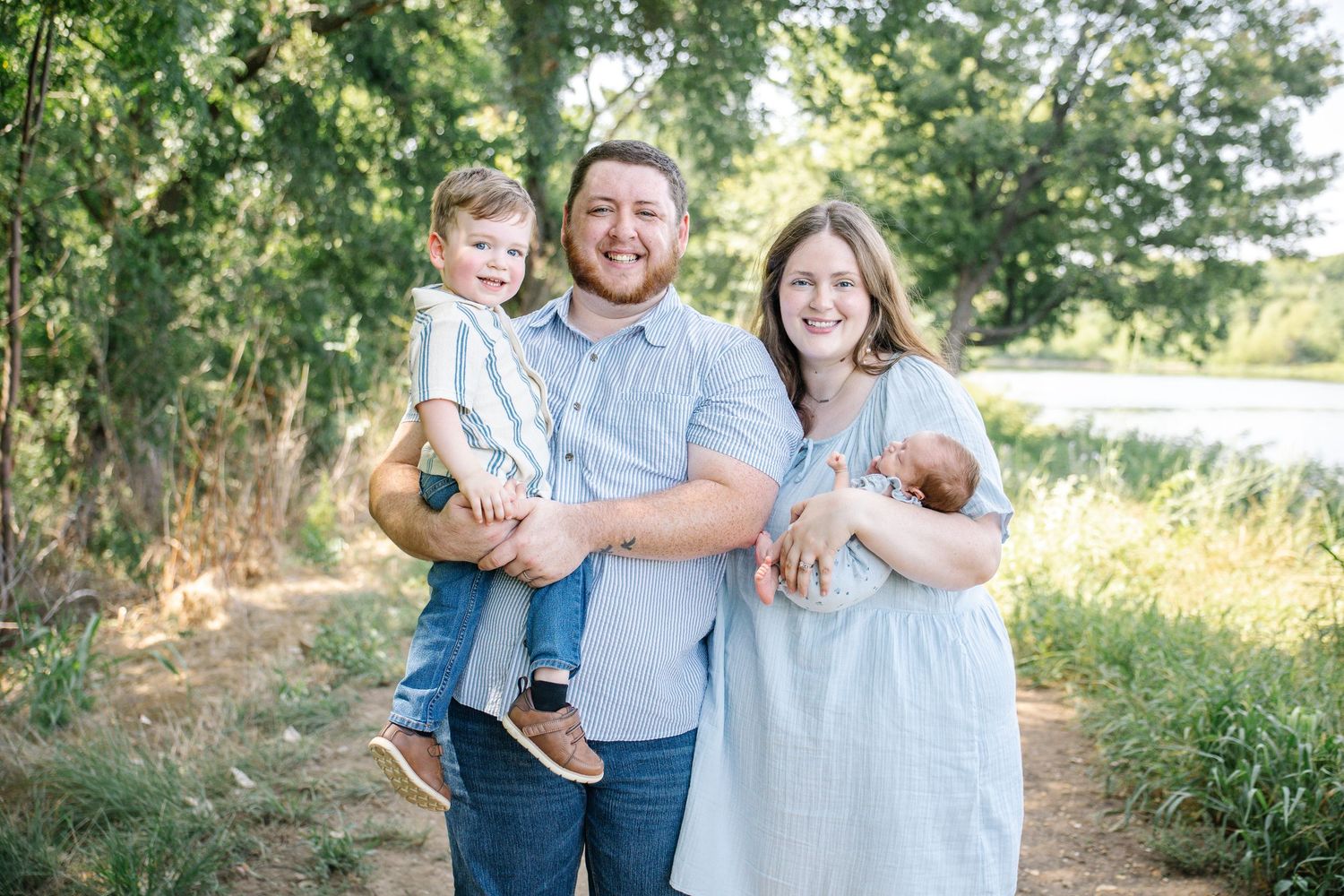 Happy family of four poses outdoors near trees and a pond, parents smiling with toddler and newborn baby.