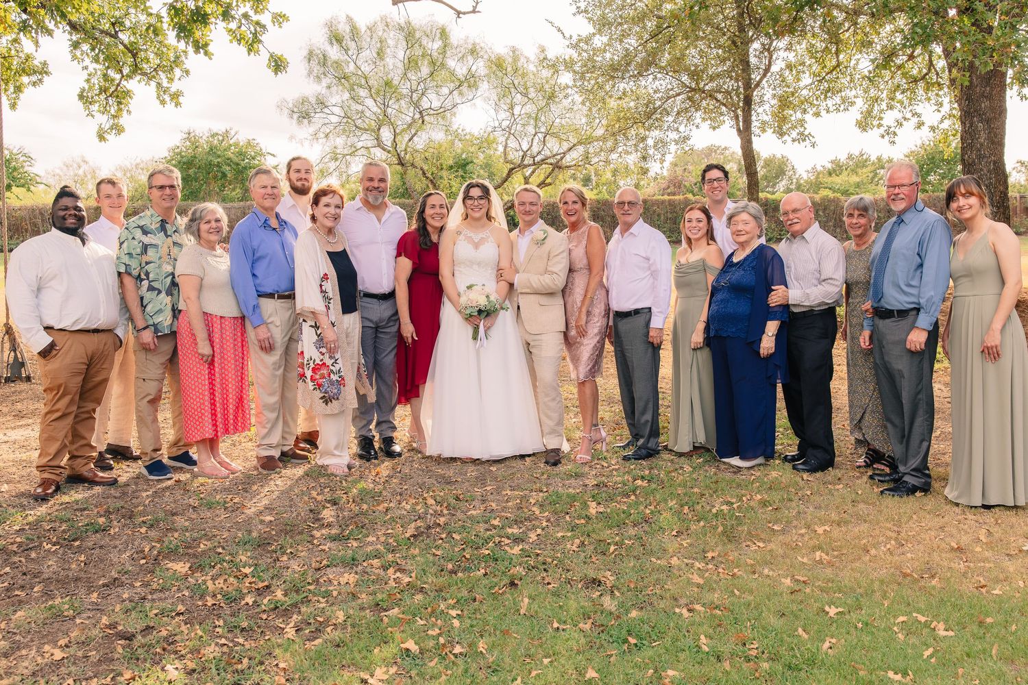 Bride and groom pose with their smiling family and wedding party in a group portrait outdoors under shady trees.