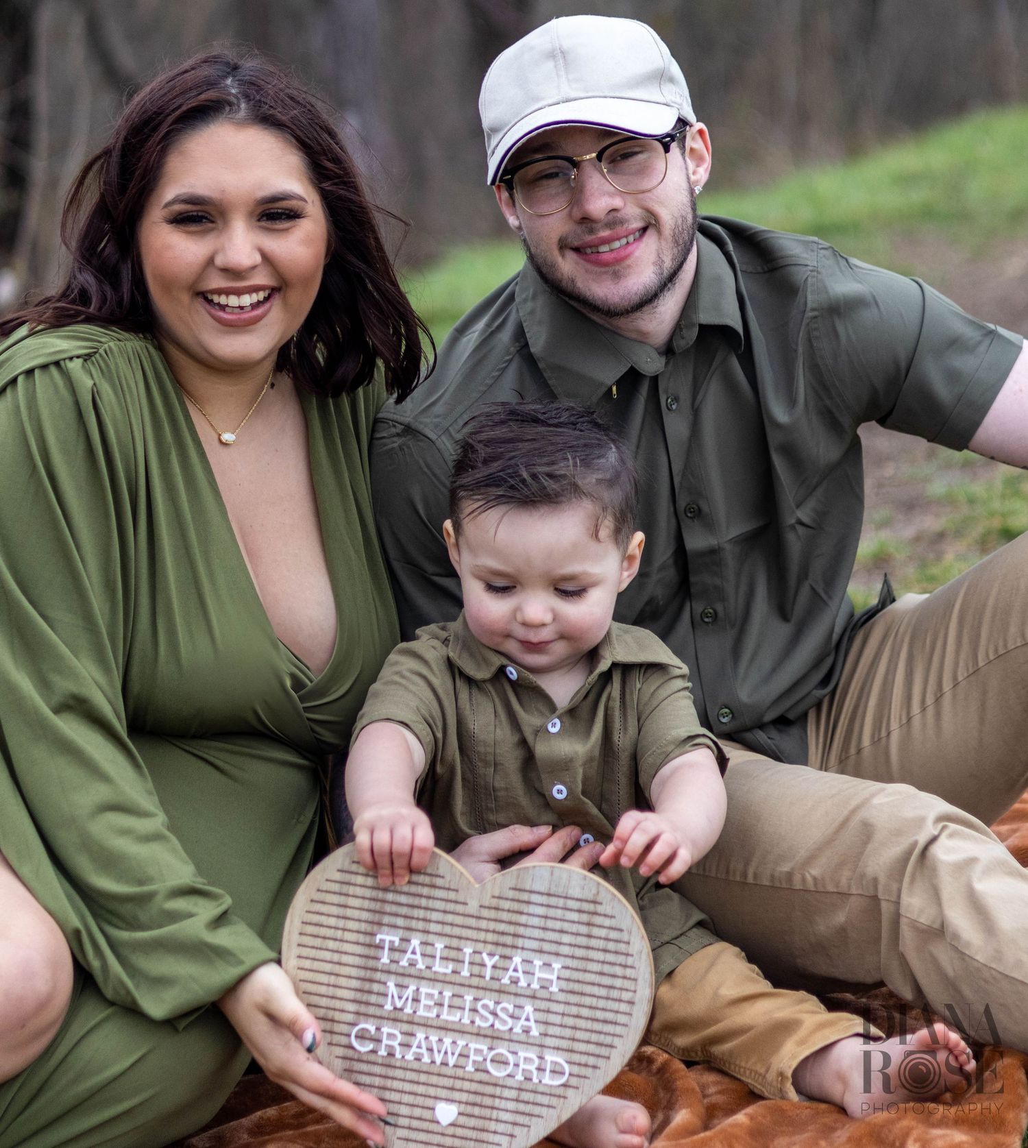 Smiling family of three holds heart-shaped letter board reading Taliyah Melissa Crawford during maternity session.